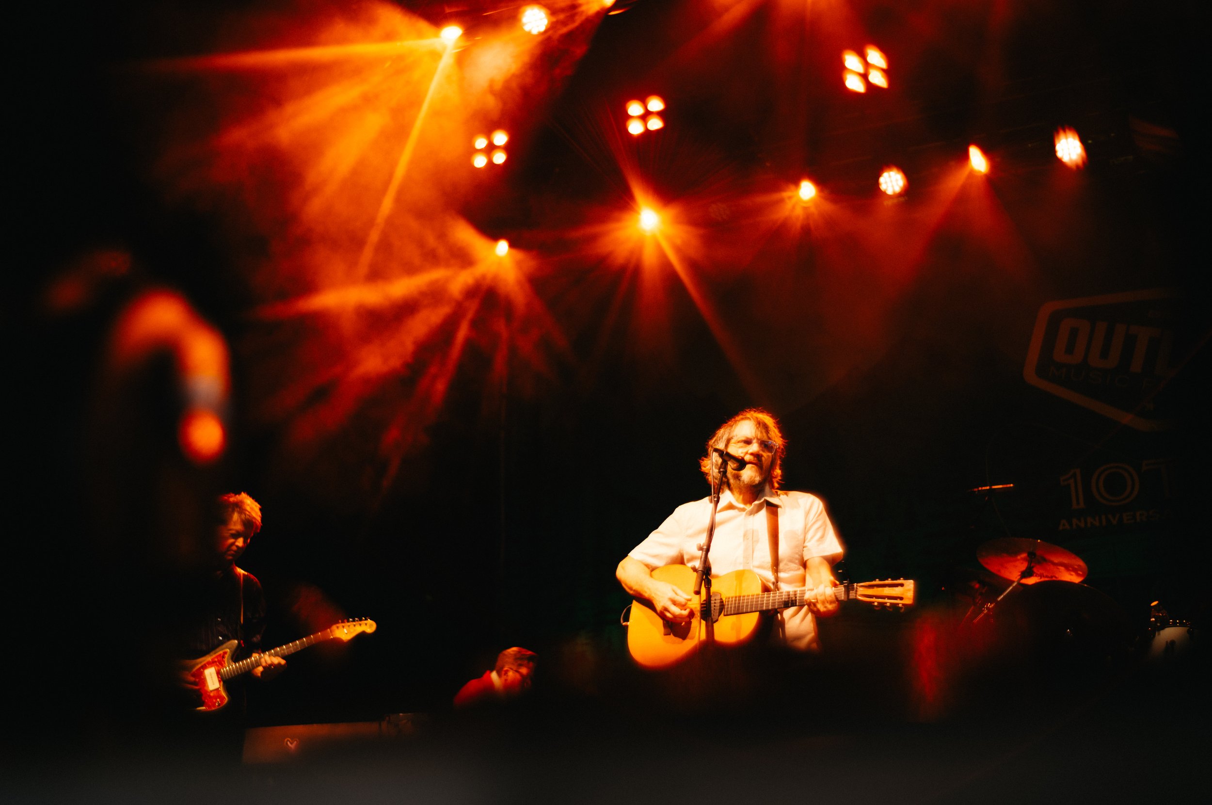 Musicians performing live on stage under red and orange stage lights, including a man with glasses playing guitar and singing, with other band members visible in the background. Jeff Tweedy of Wilco playing at The Outlaw Festival.