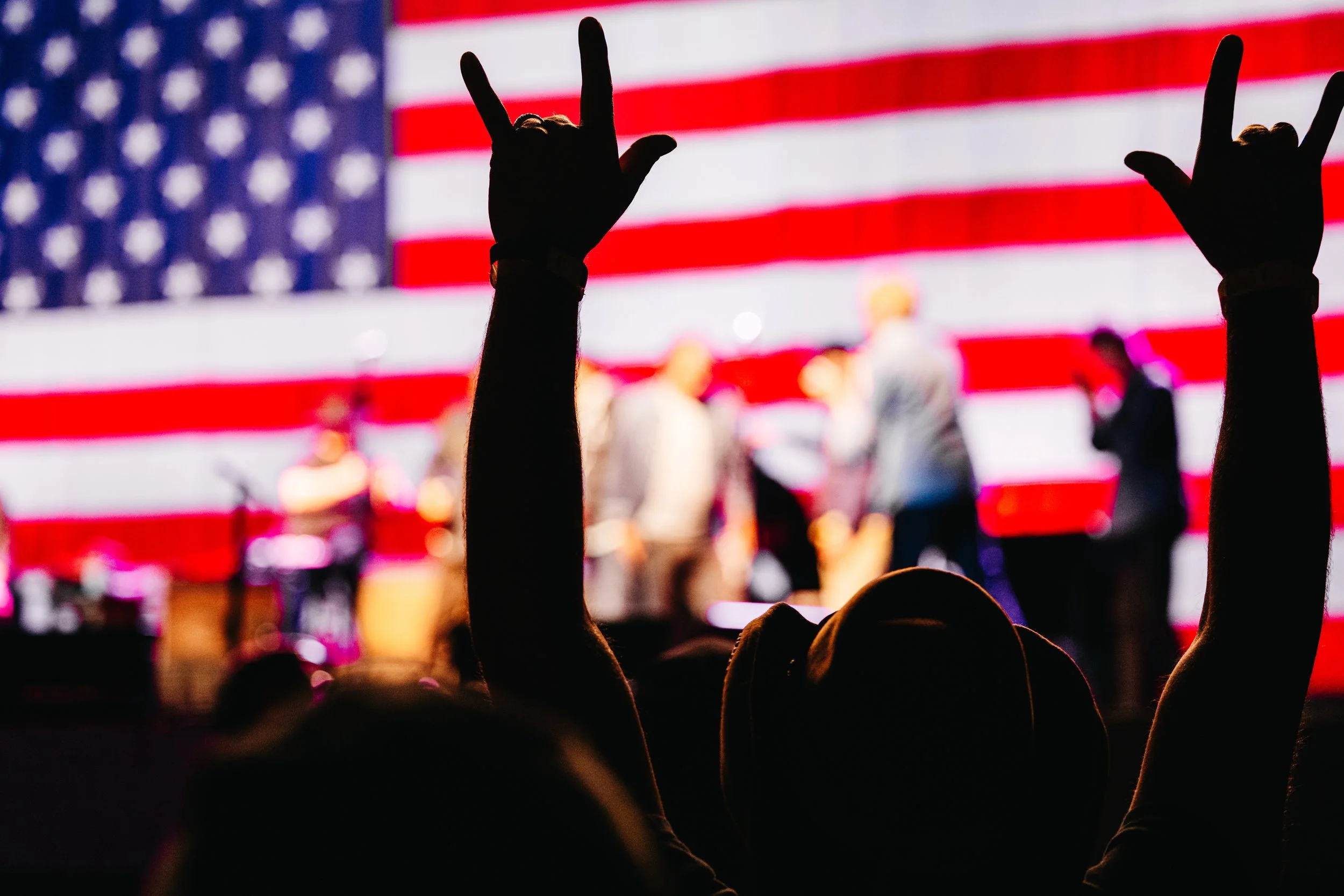Silhouettes of hands making rock and peace signs with an American flag in the background at a crowded event.