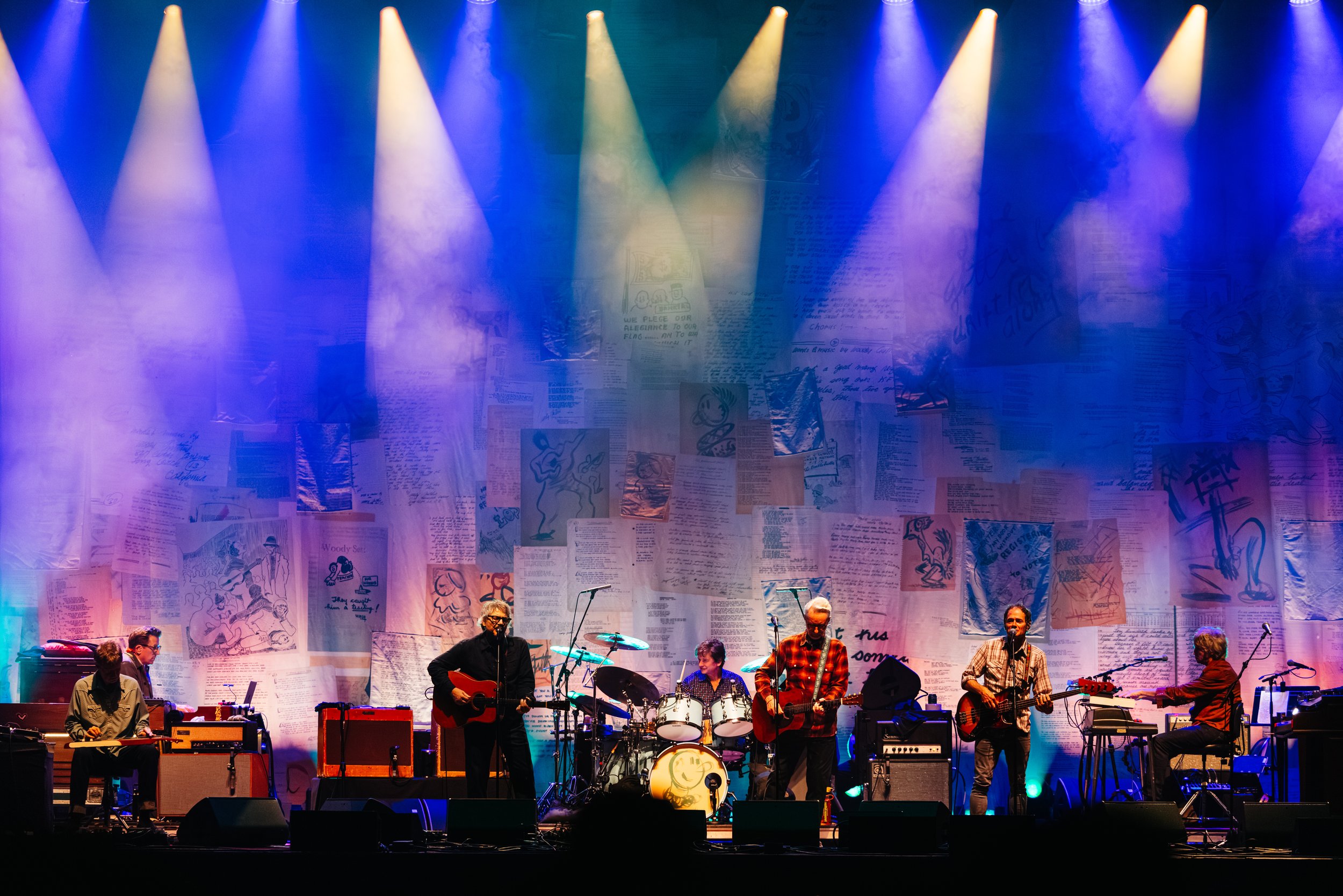 Musicians performing on stage with a colorful backdrop of drawings and text, illuminated by stage lights.