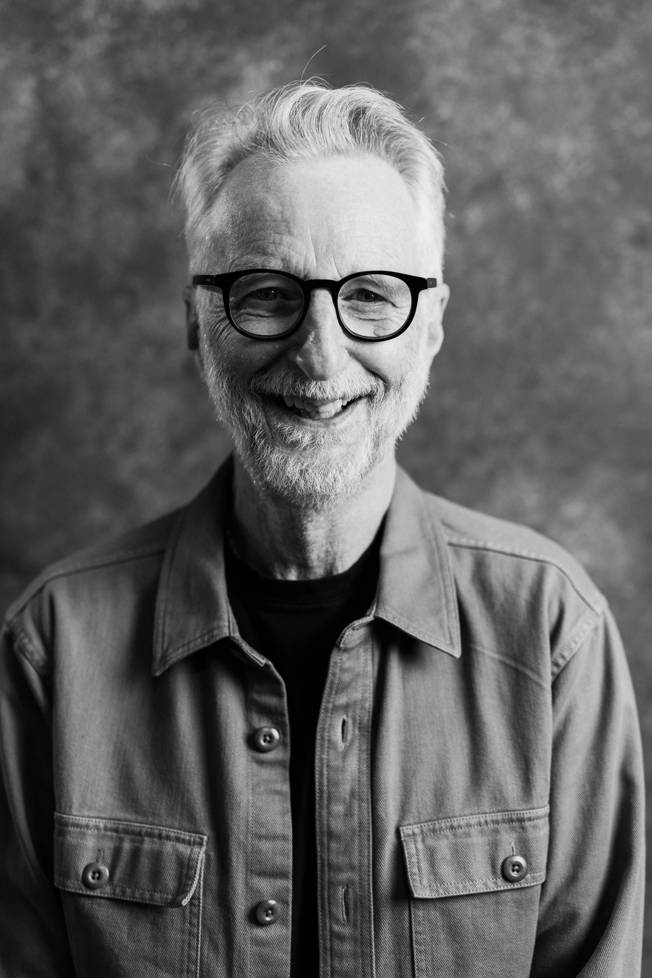 Black and white photo of an elderly man with gray hair, beard, and glasses, smiling and wearing a buttoned denim shirt. Billy Bragg Portrait.