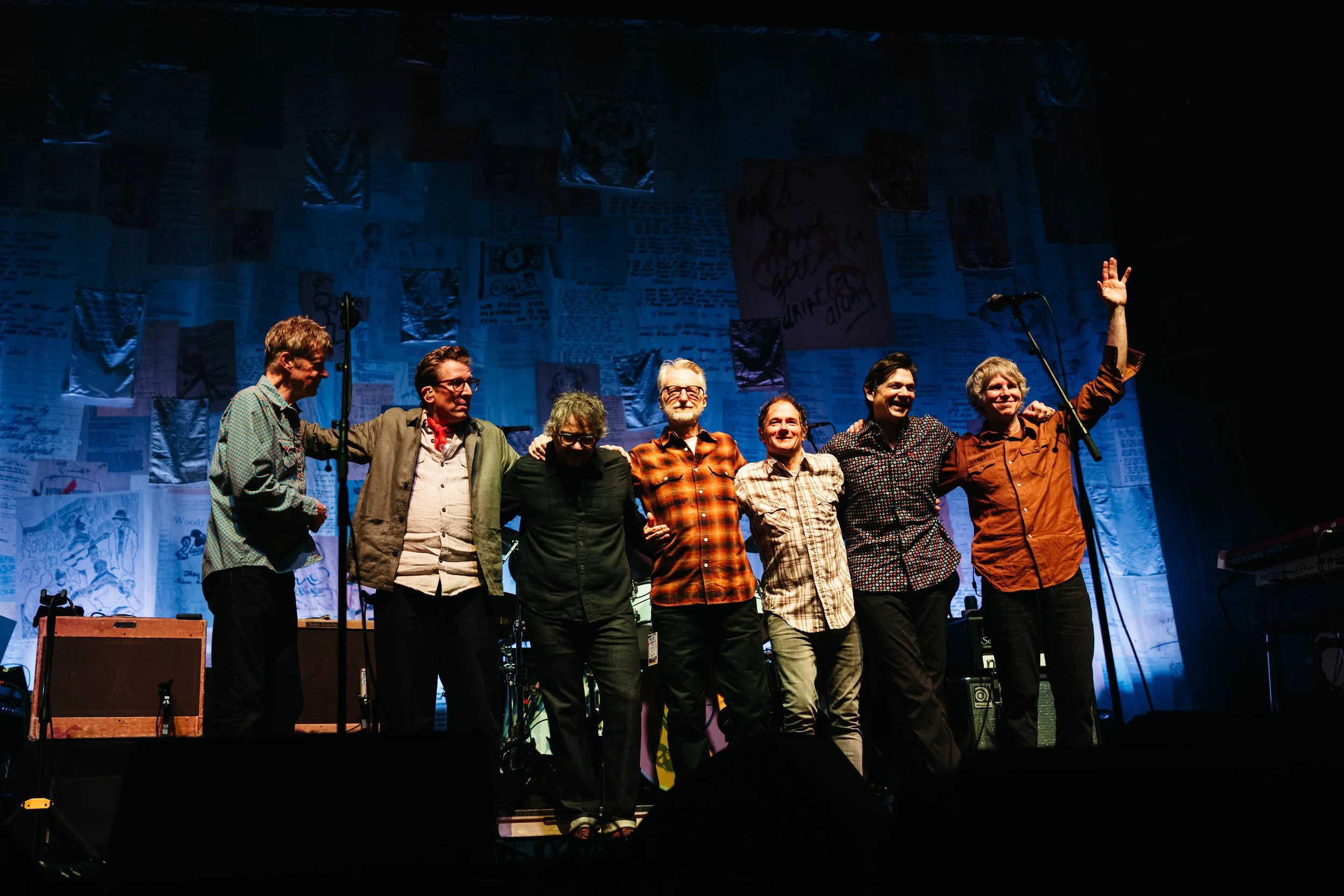 A group of seven people standing on a stage, smiling and embracing each other, with a dark background and various papers and posters hanging overhead. One person is raising their hand.