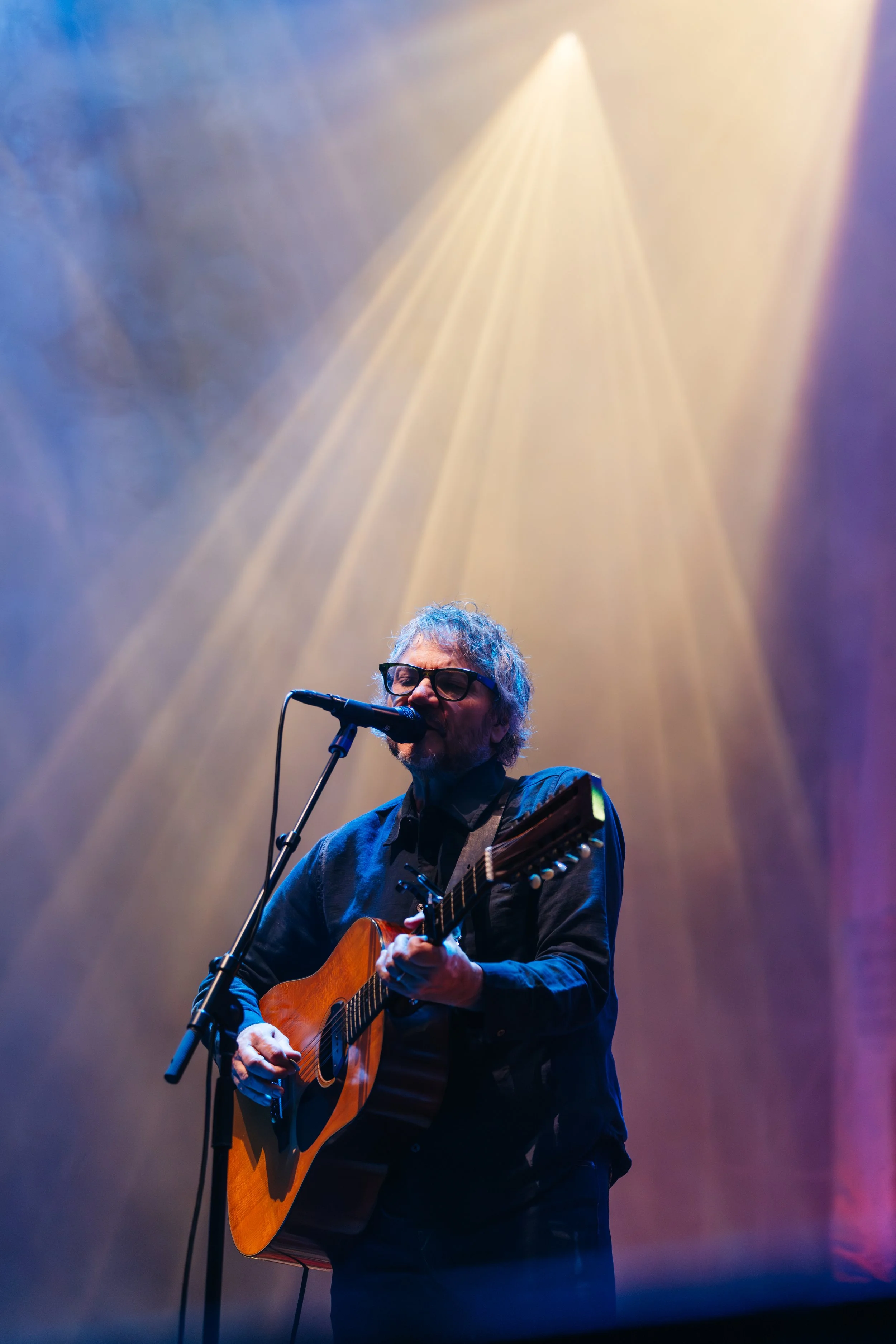 An older man with glasses, gray curly hair, and a beard is playing an acoustic guitar and singing into a microphone on stage, with colorful stage lights shining down.