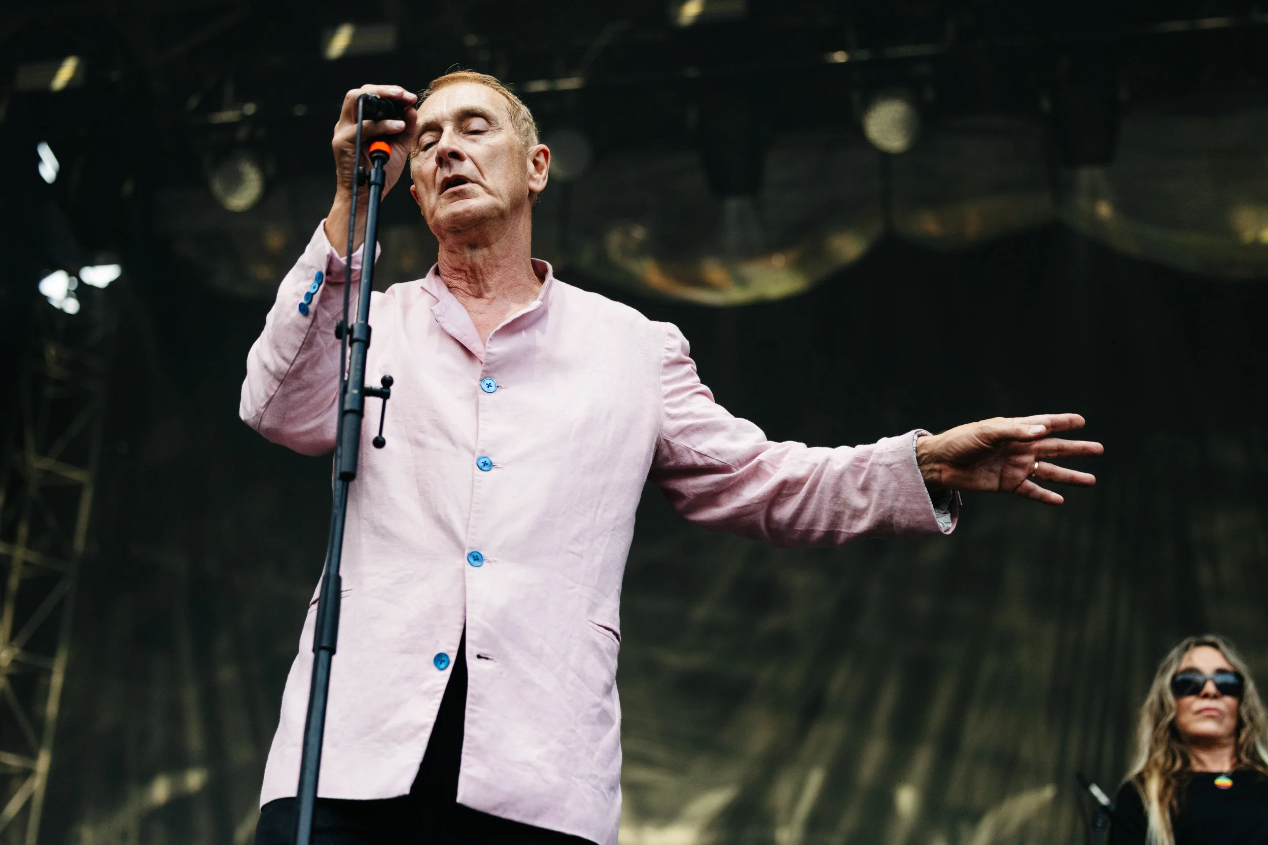 A man in a pink shirt singing or speaking into a microphone on a stage with a dark background.