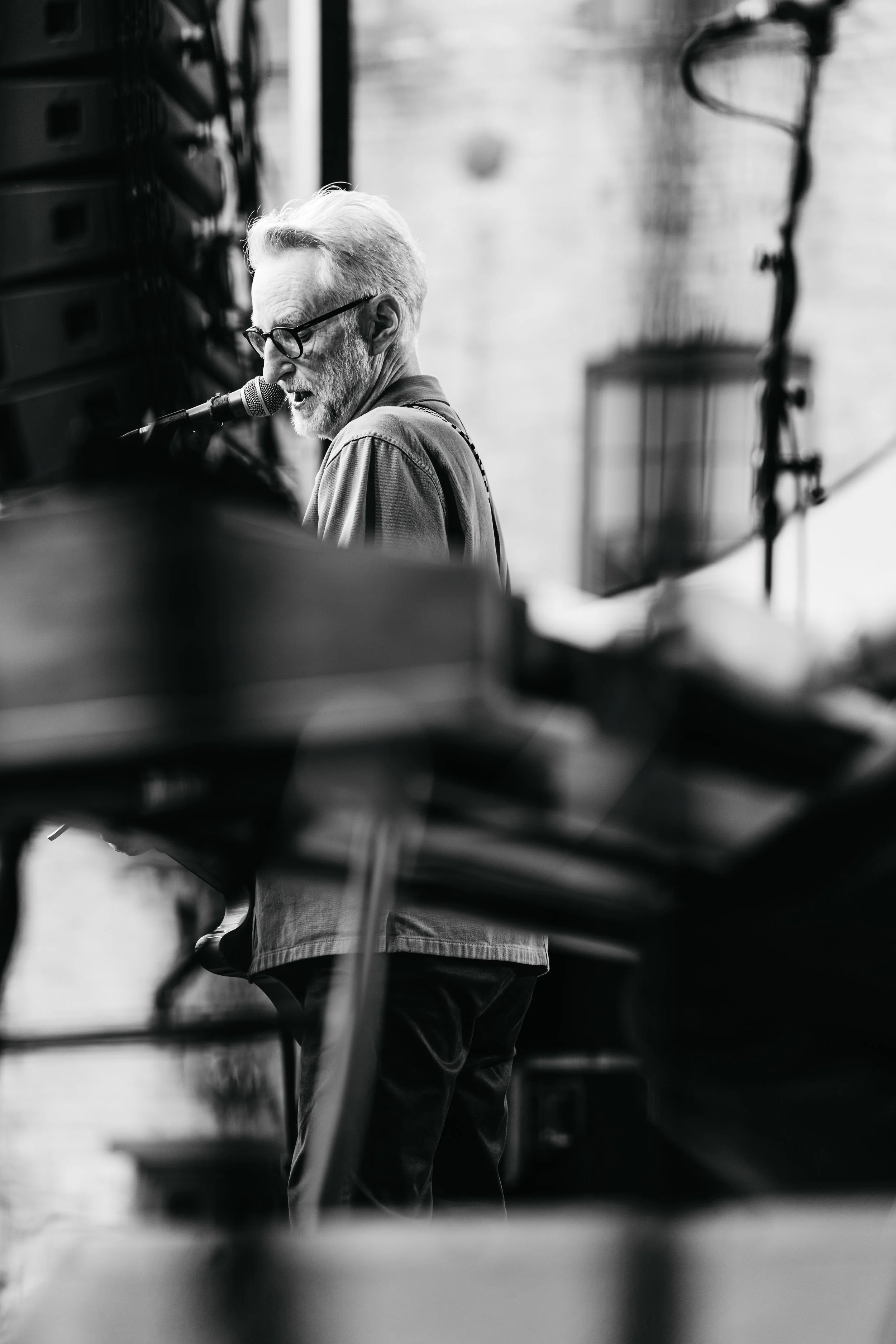 A black-and-white photograph of an elderly man with glasses and a beard, playing a piano and singing into a microphone. The background shows a blurred wall and some equipment.