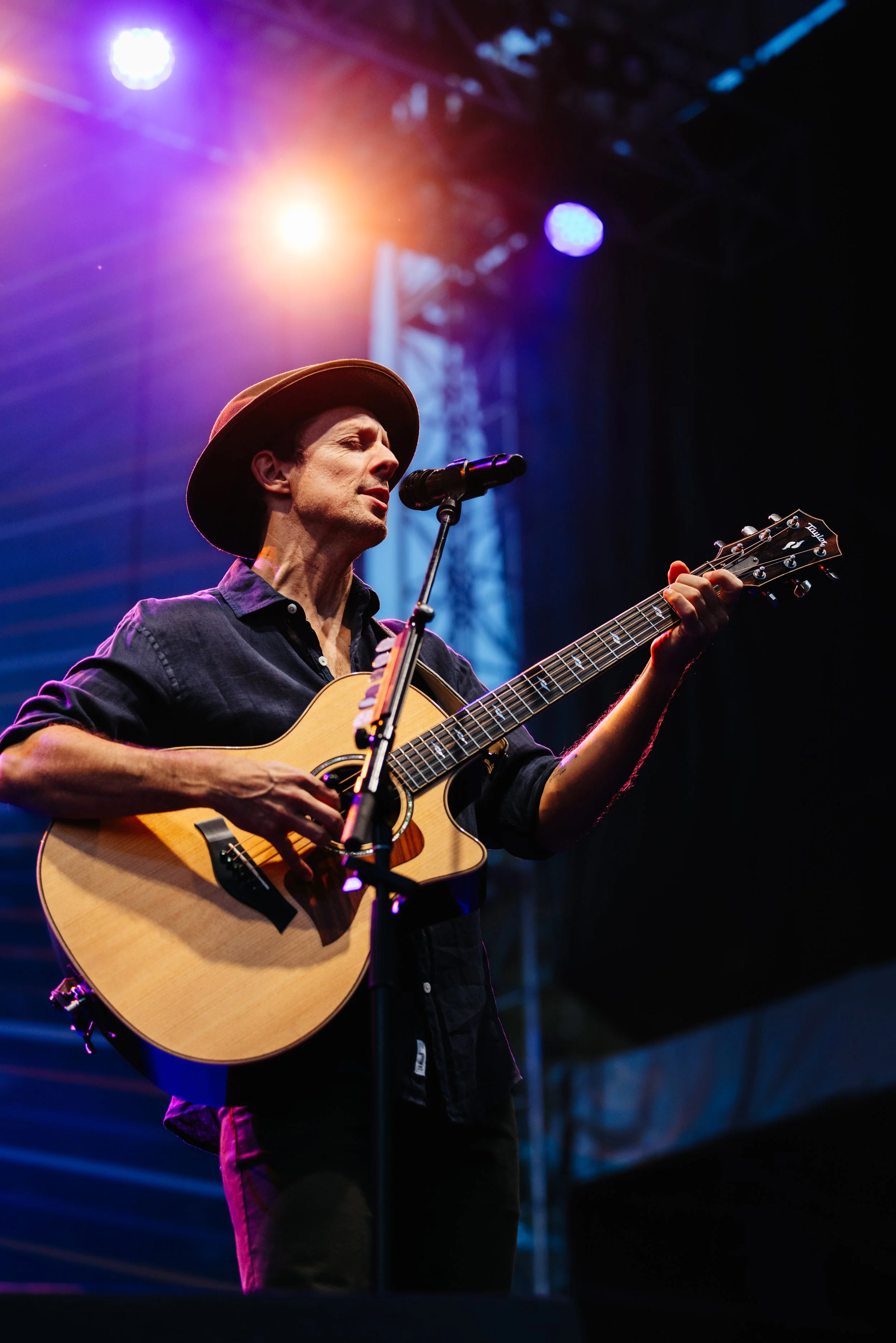 A man wearing a hat and dark clothing is playing an acoustic guitar and singing into a microphone on a stage illuminated by bright colored stage lights. Jason Mraz.