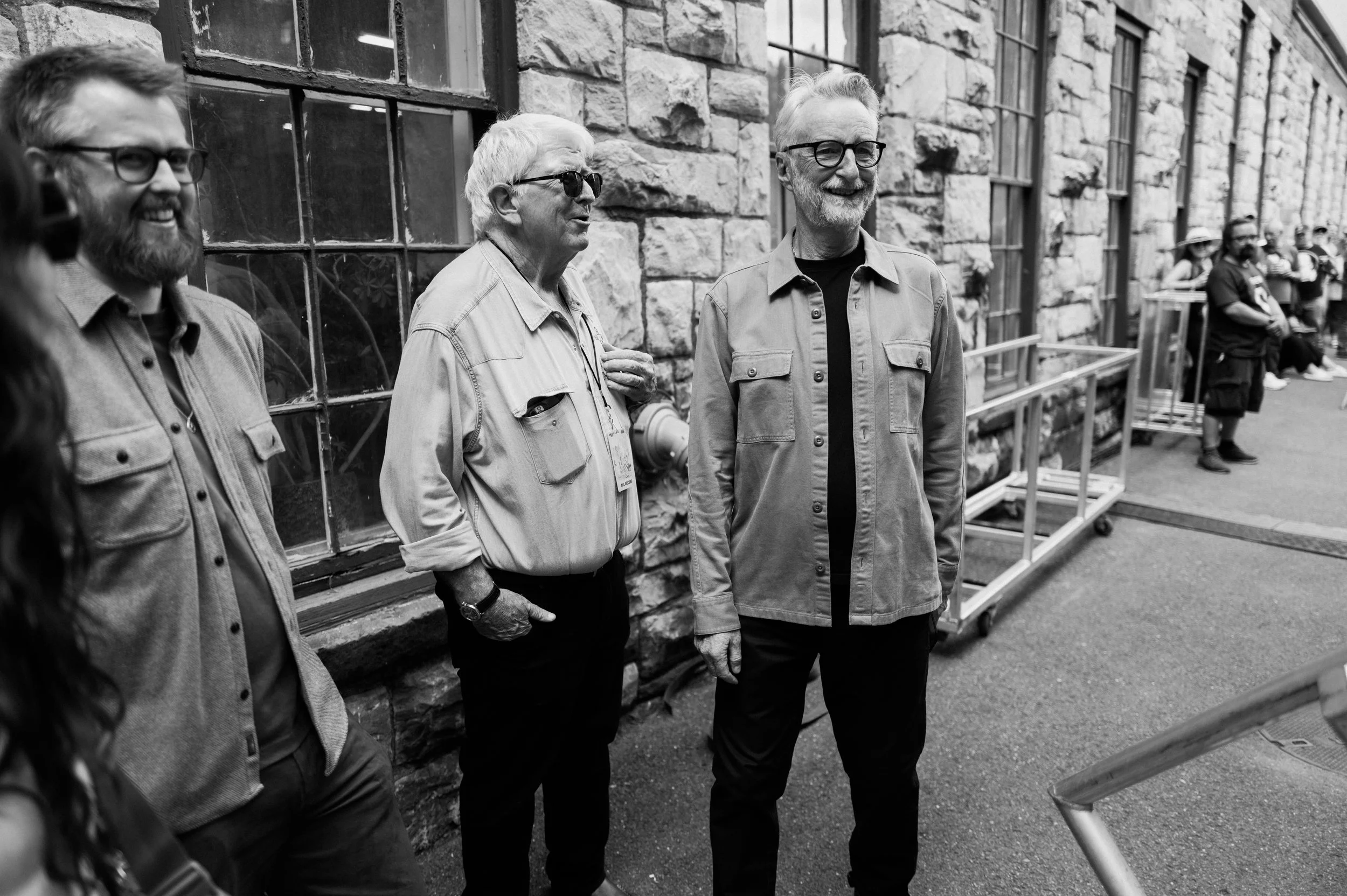 Four men standing outside near a stone building with large windows, engaged in conversation and smiling. Billy Bragg.
