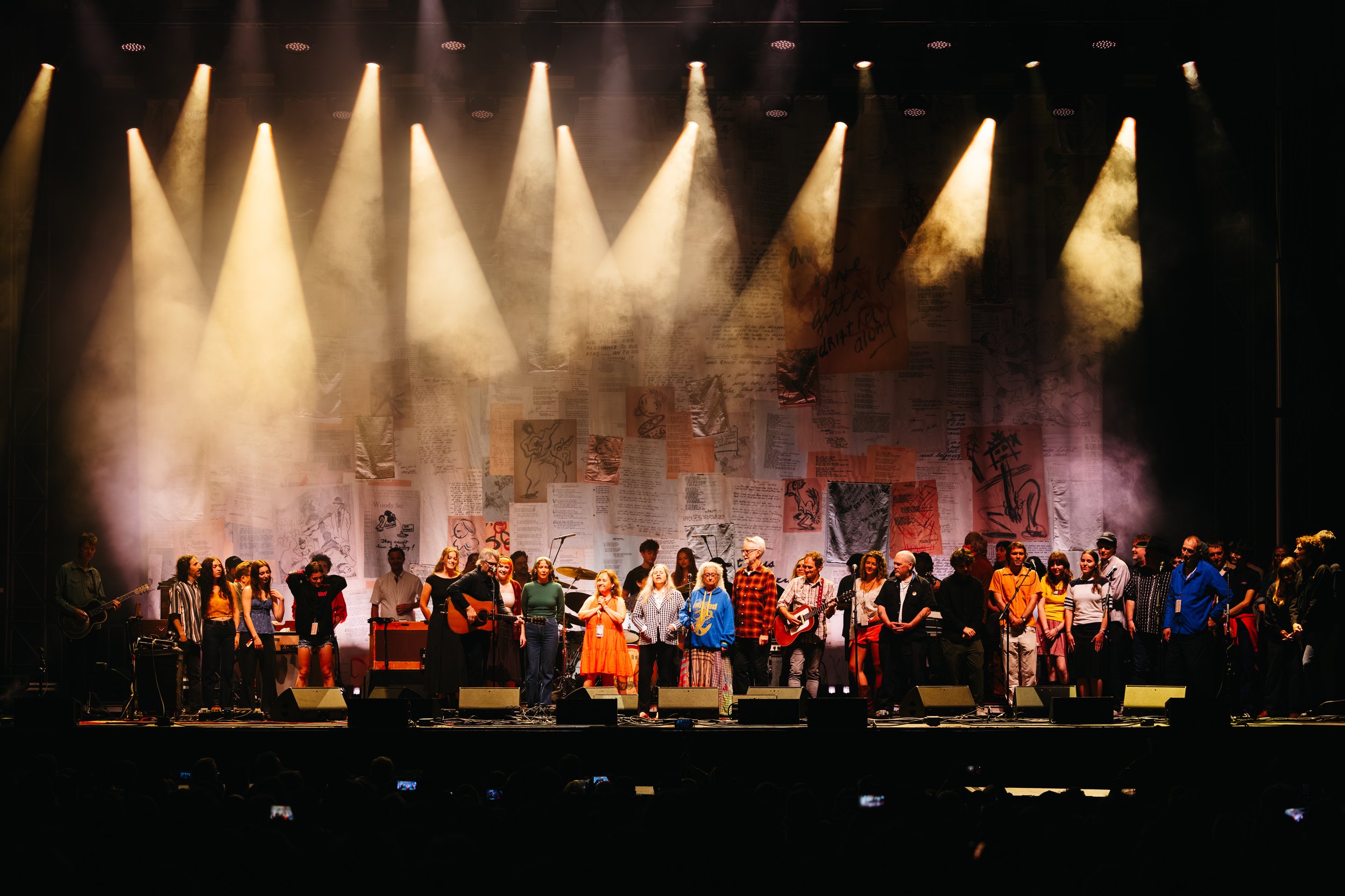 A large group of people on stage during a performance or rehearsal, with stage lights shining down, and a backdrop of various posters and artwork.