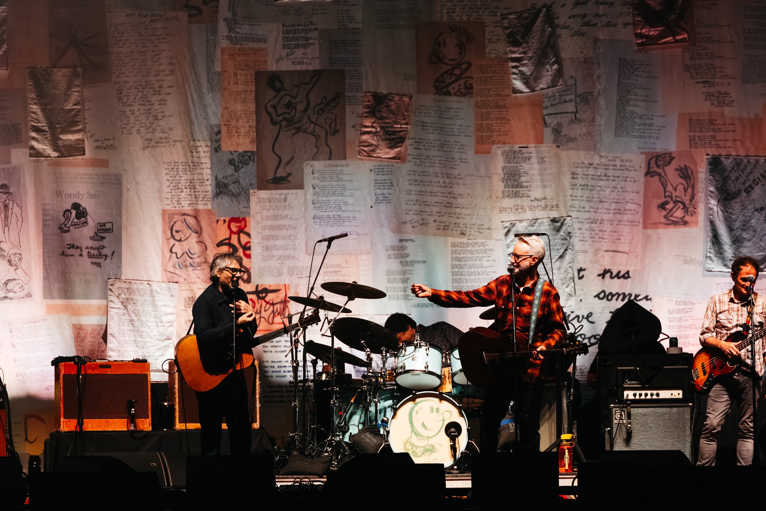 A band performing on a stage with a backdrop made of various handwritten and illustrated papers. The band members are playing guitars and drums, and are engaged in their performance.