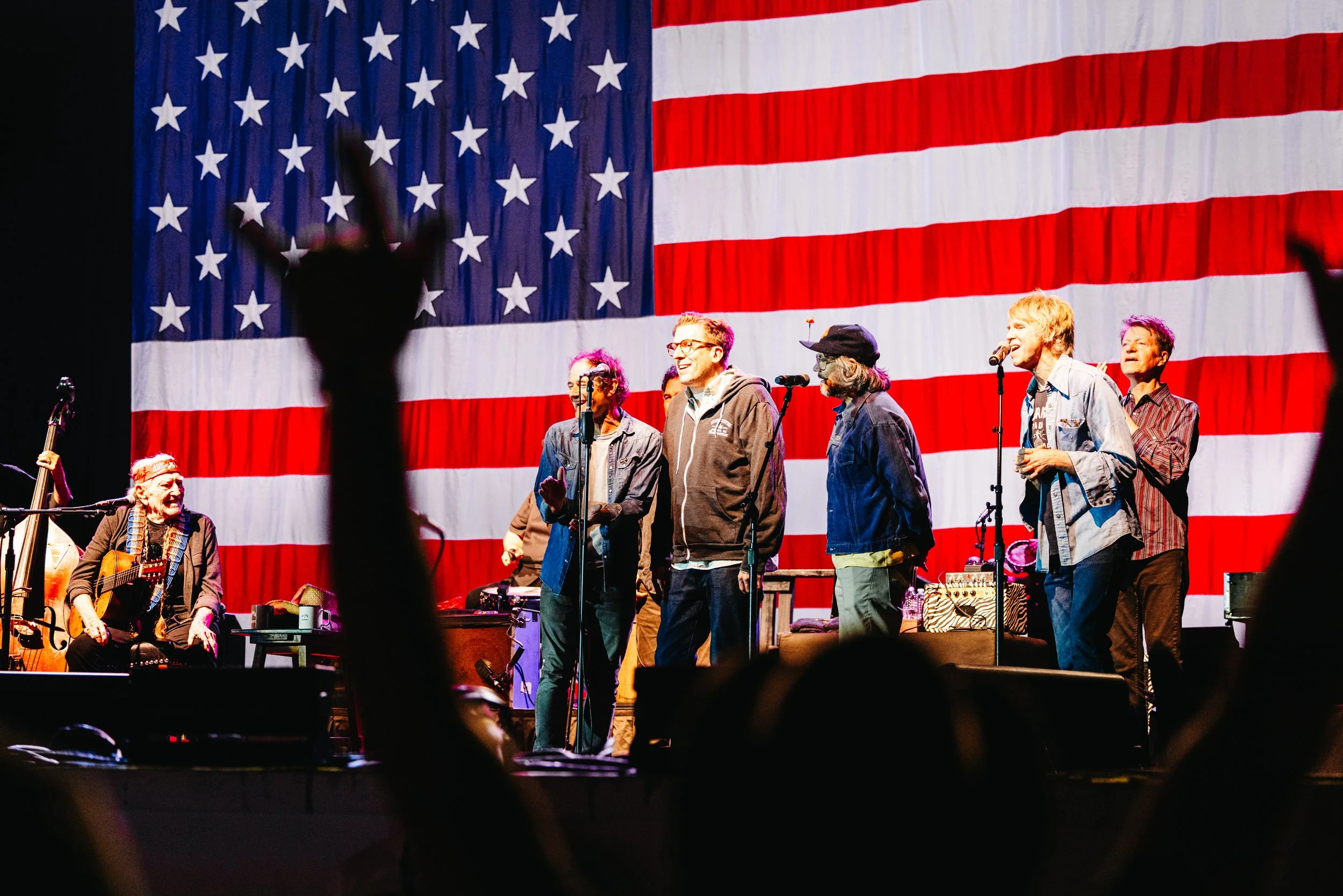 A group of performers on stage in front of an American flag backdrop, with some holding microphones and others playing instruments.