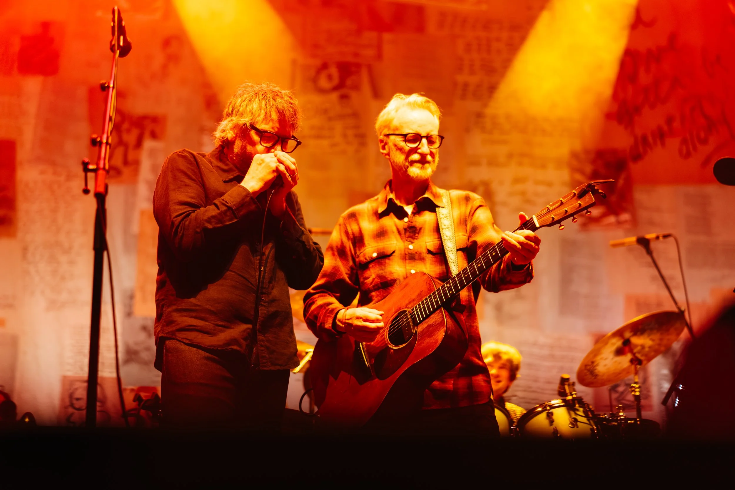 Two men performing music on stage, one singing into a microphone and the other playing an acoustic guitar, illuminated by warm orange stage lights.