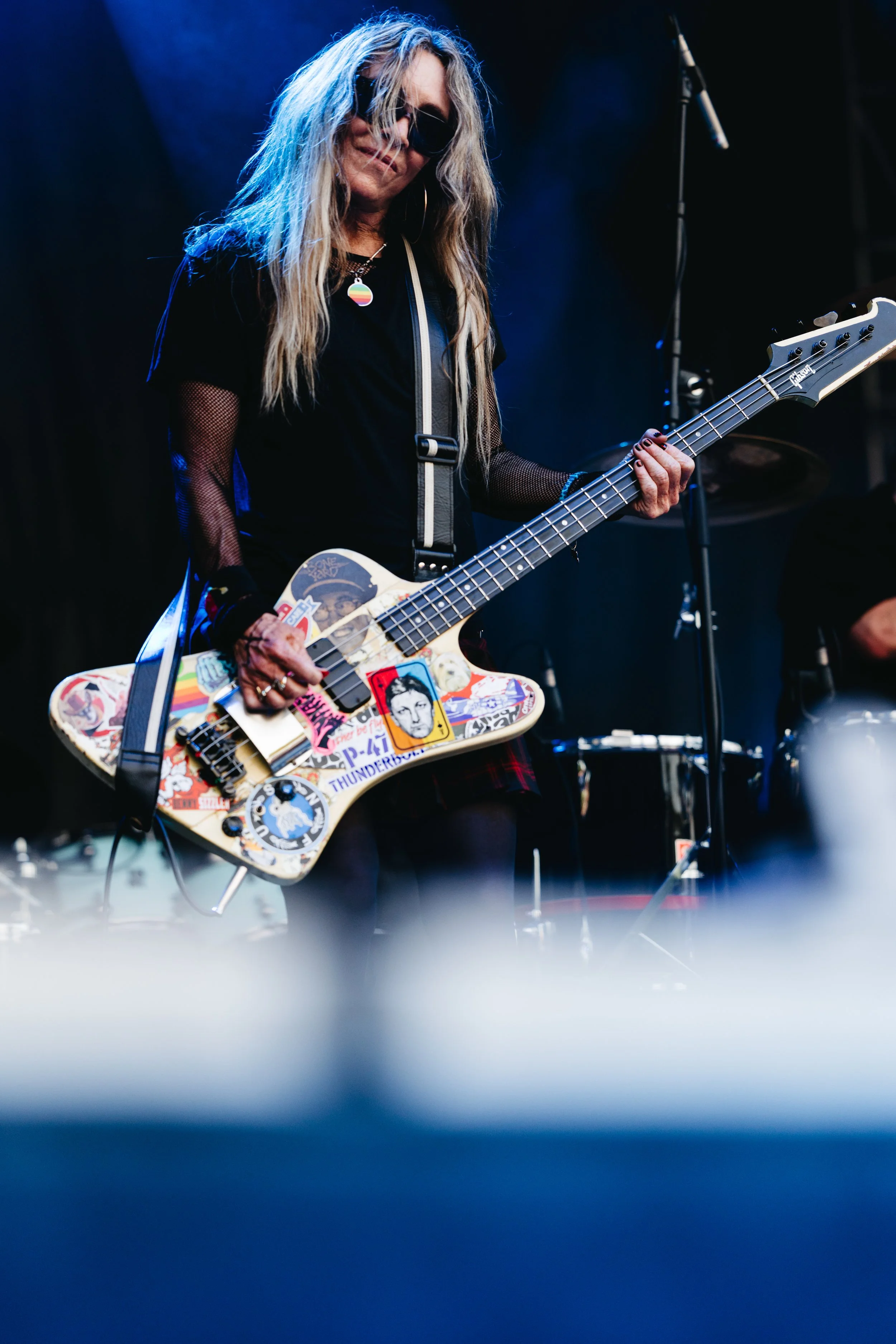 A woman with long wavy blonde hair wearing sunglasses and a black mesh top playing a sticker-covered bass guitar on stage.