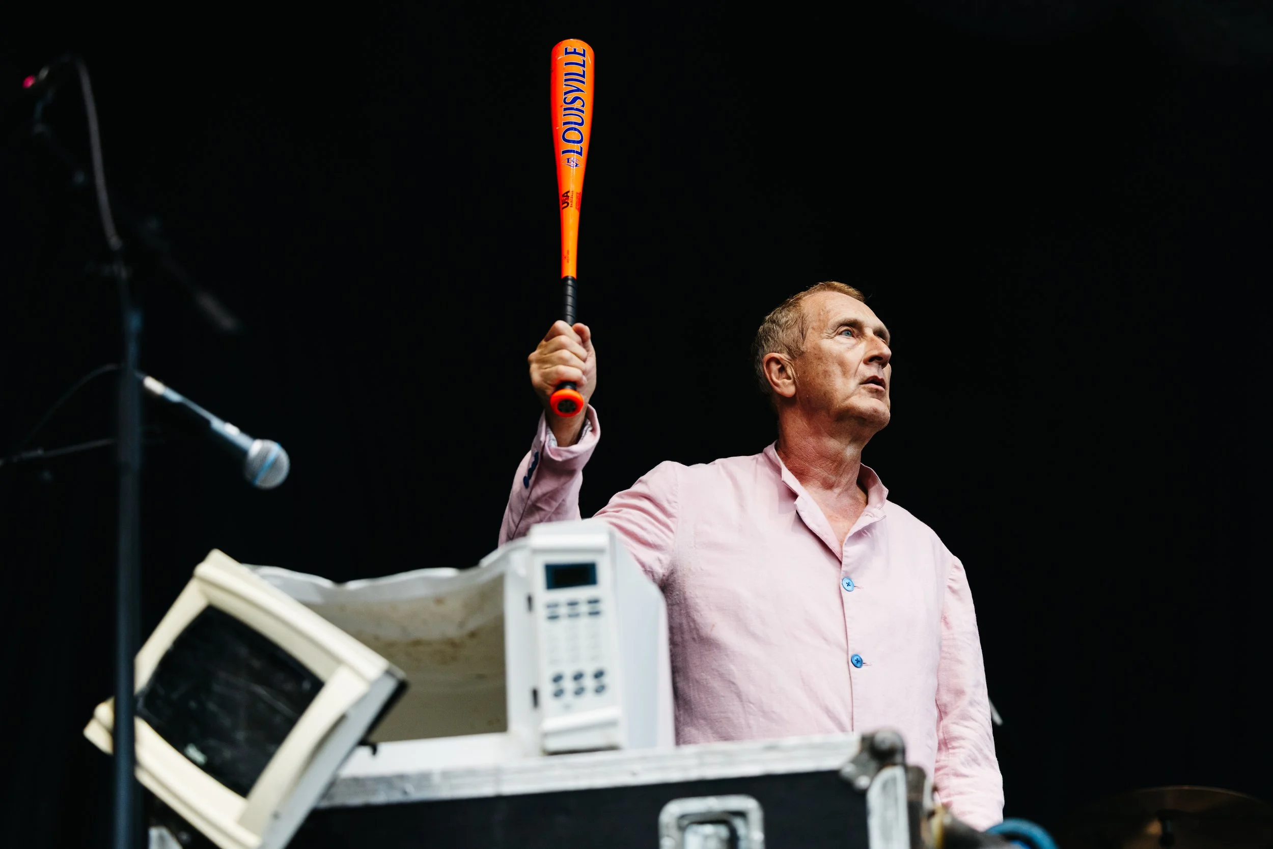 A man in a pink shirt holding a Louisville baseball bat, standing indoors with various electronic devices and a microphone nearby.