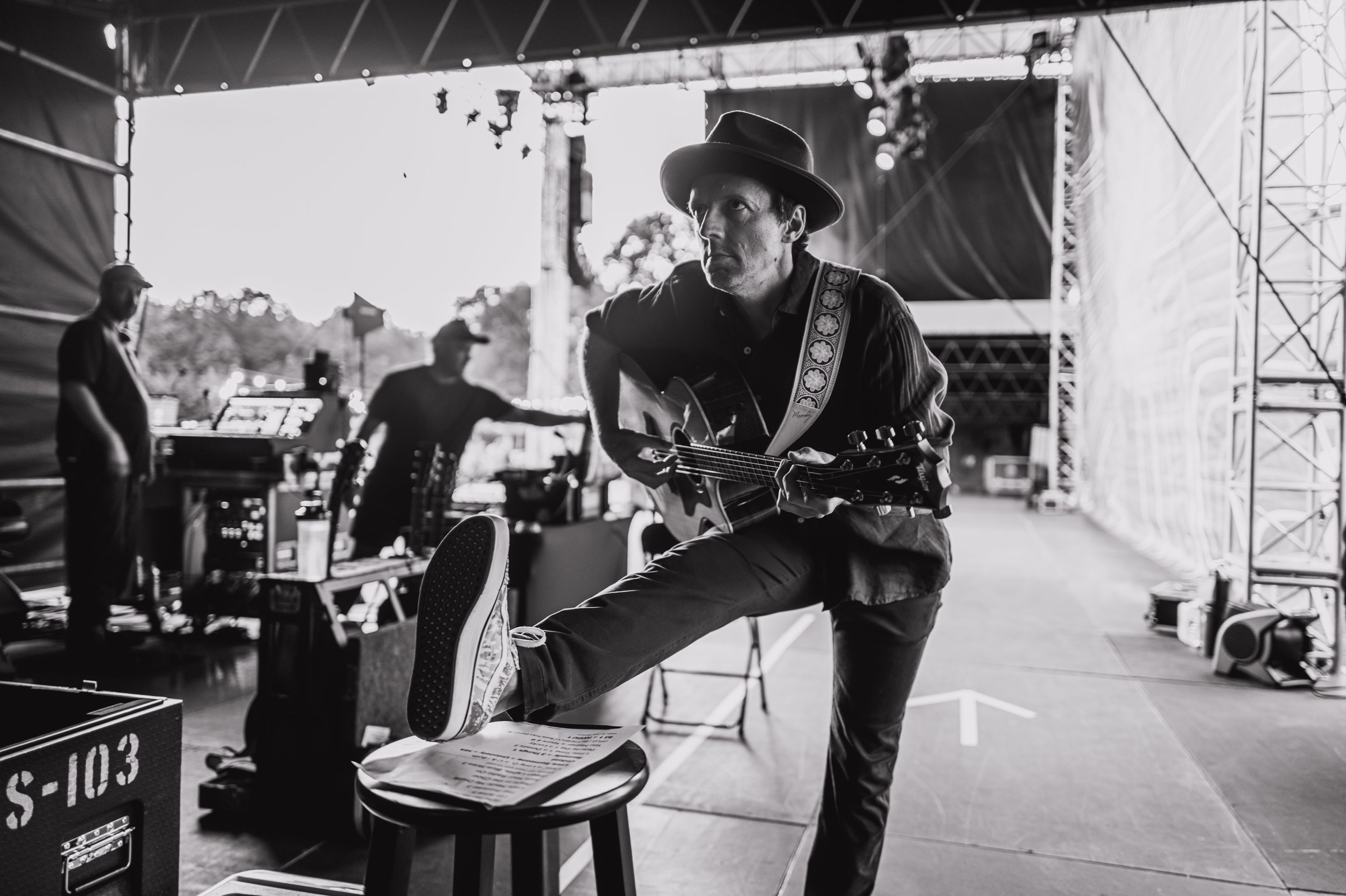 Black and white photo of a man wearing a hat playing a guitar on a stage. Two other men are standing in the background near audio equipment. The setting appears to be an outdoor event under a large tent. Jason Mraz.