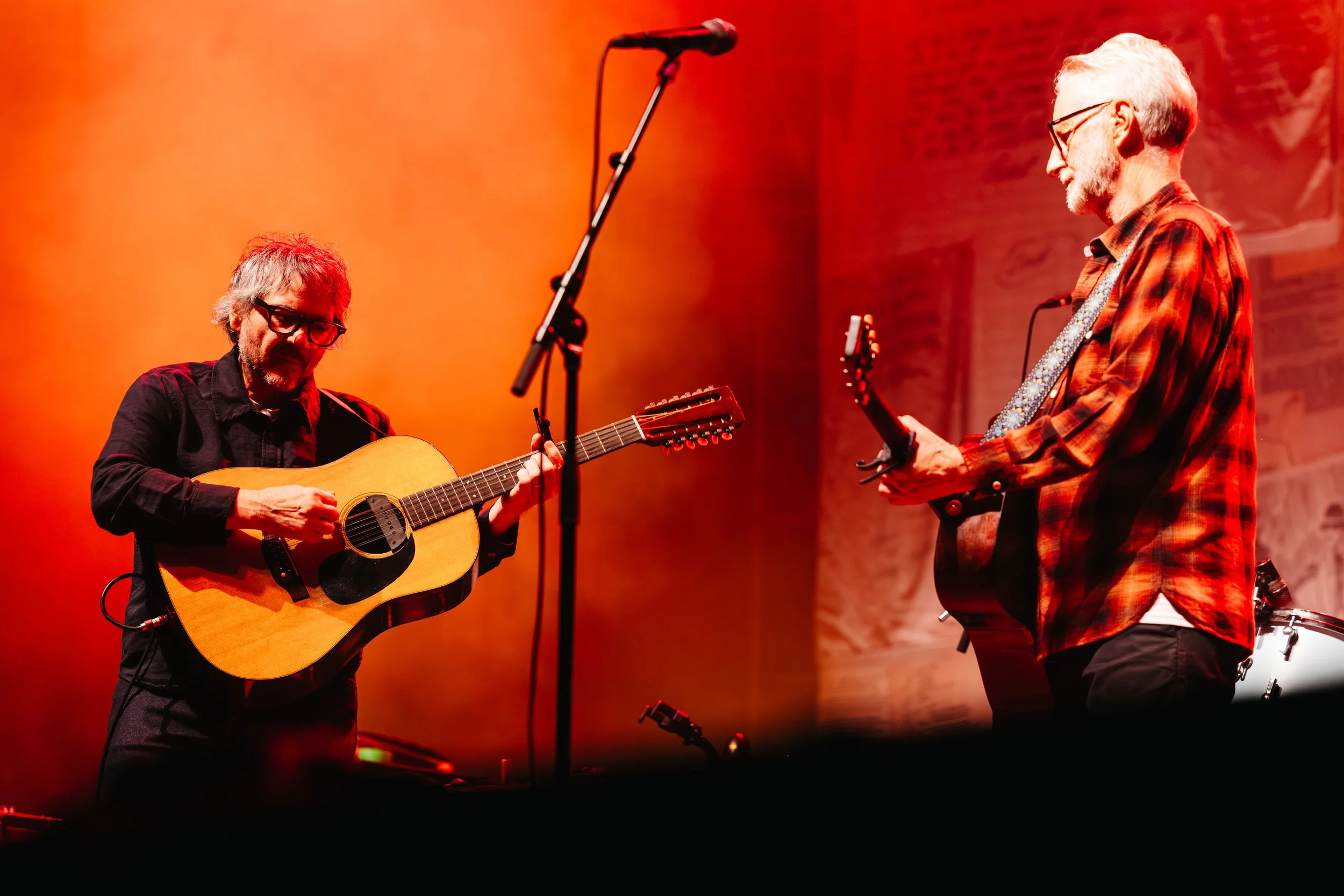 Two musicians performing on stage: one playing an acoustic guitar and the other holding an electric guitar, with orange stage lighting in the background.