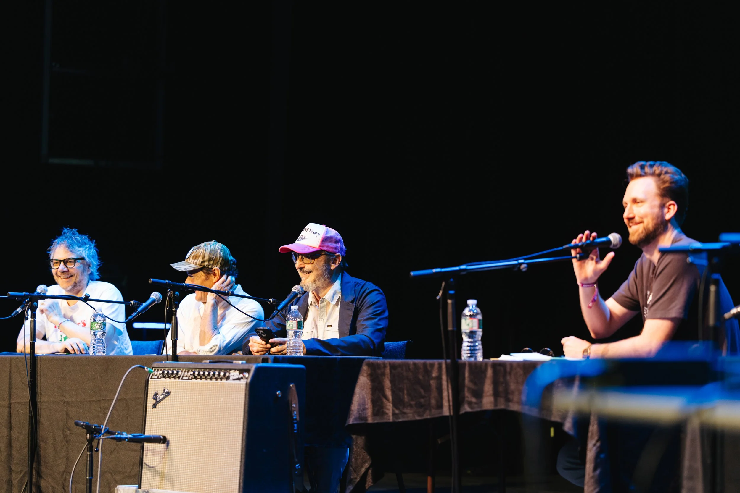 Four men sitting at a panel discussion on stage, with microphones and water bottles in front of them. They are smiling and appear to be enjoying the event.