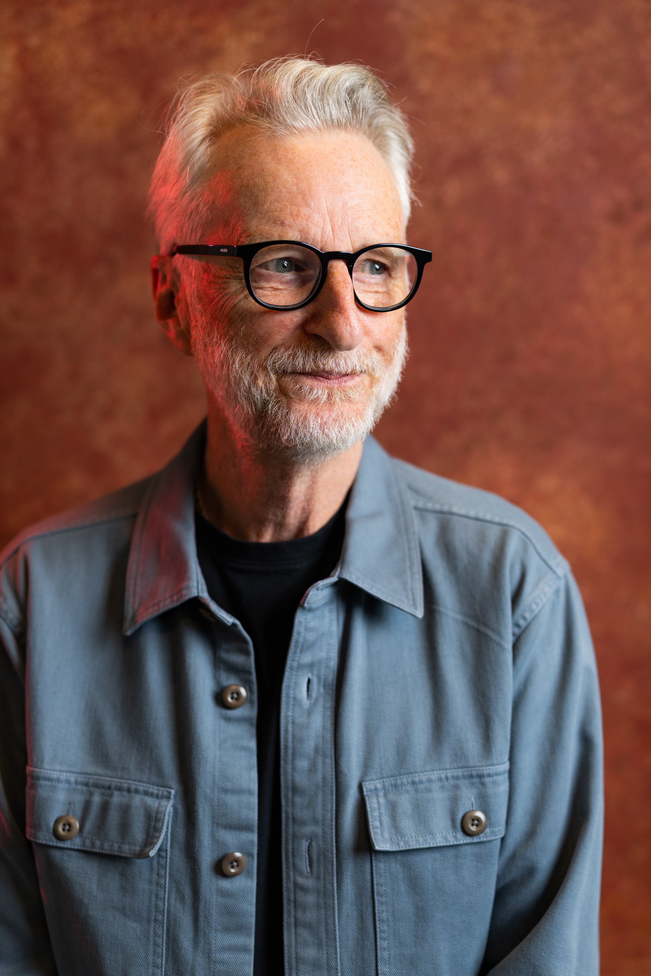 An older man with gray hair, beard, and glasses, smiling slightly, wearing a denim jacket over a black shirt, with a warm, textured background. Billy Bragg.
