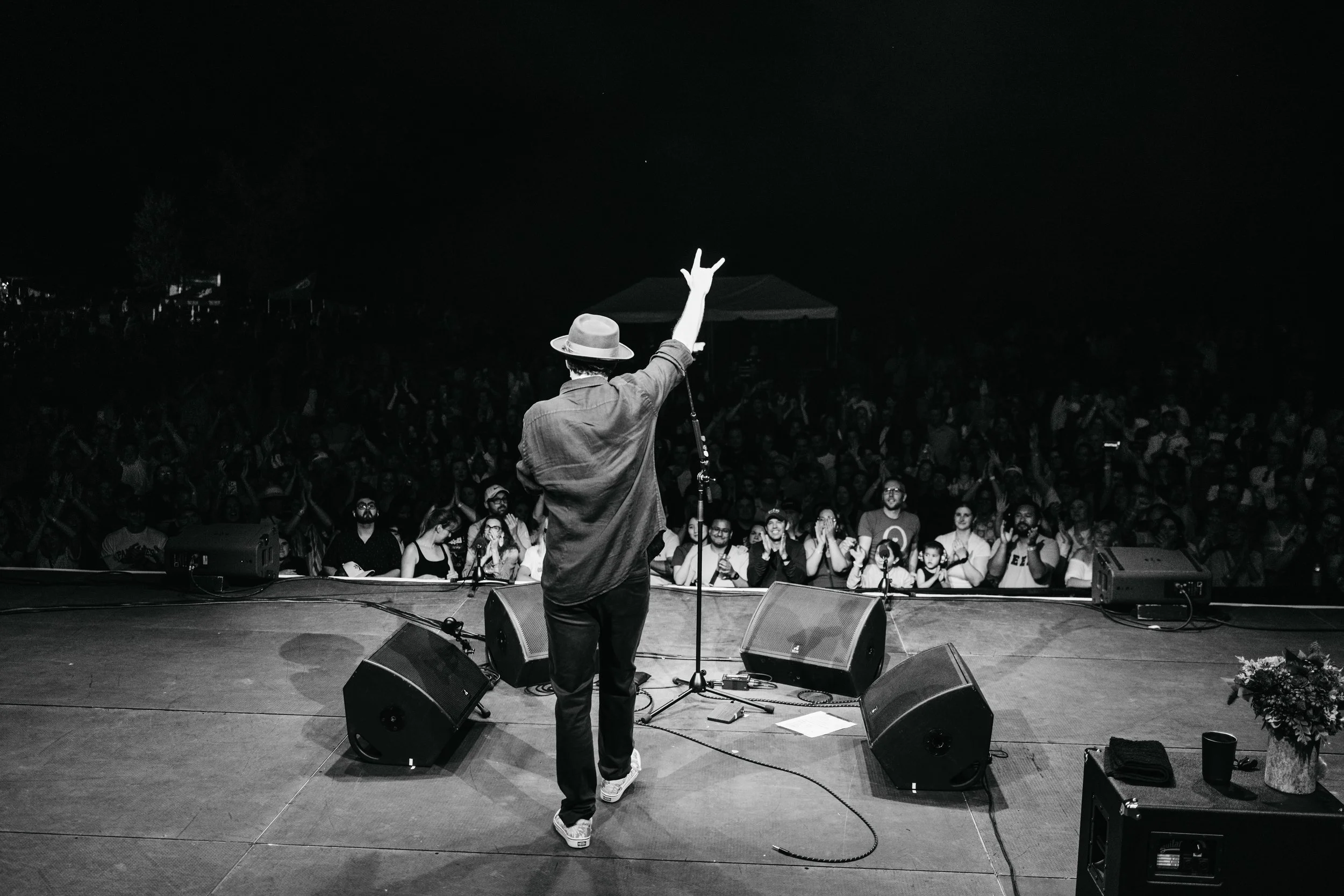 A person on stage at a concert or performance, facing a large audience. The person is wearing a hat and casual clothing, holding a microphone and making a peace sign with their right hand. The audience appears engaged and excited, and the scene is in