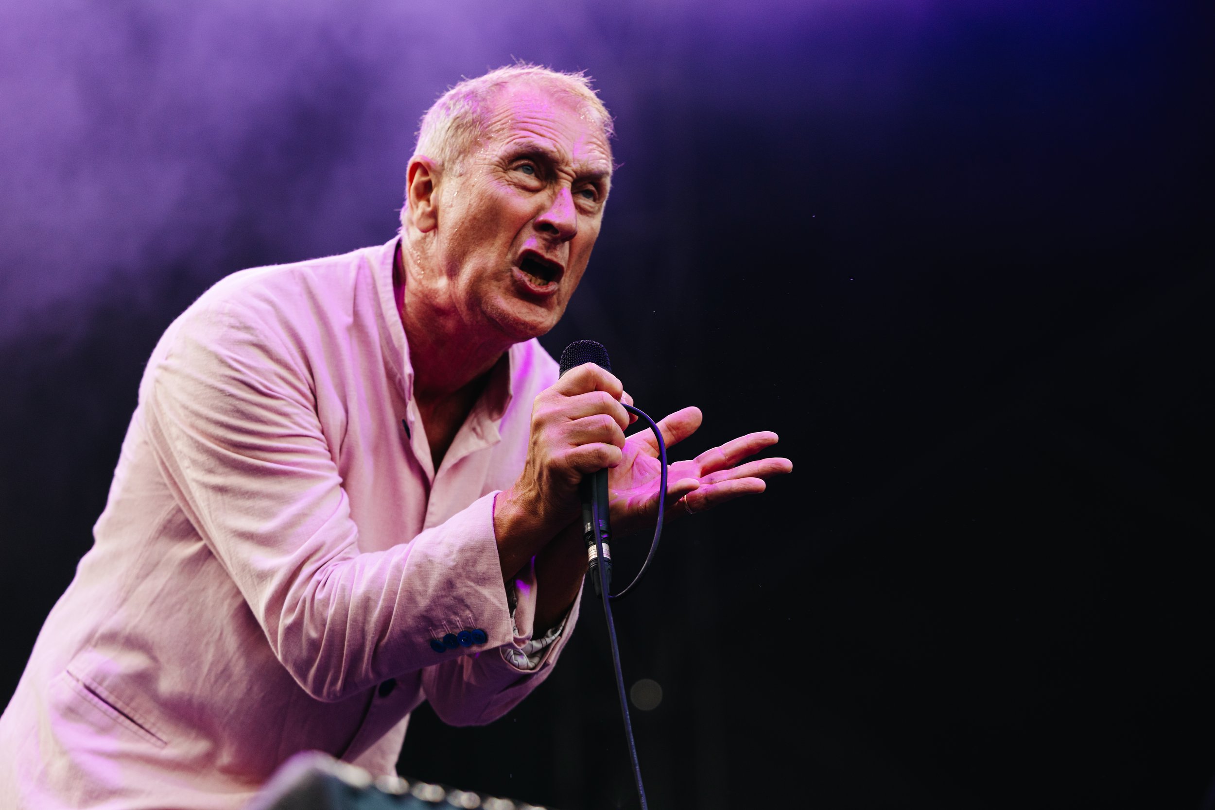 An elderly man with short, light-colored hair, wearing a light pink shirt, passionately speaking or singing into a microphone against a dark background with purple lighting.