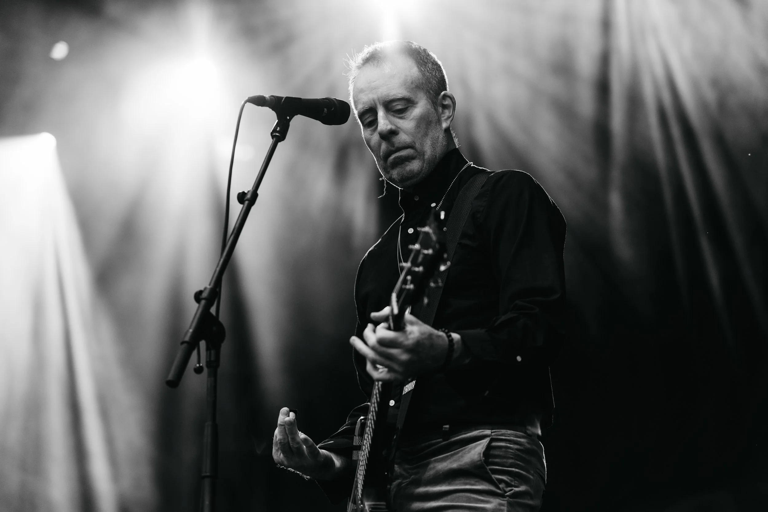 A black and white photo of a man playing guitar and singing into a microphone with his eyes closed, illuminated by stage lights.