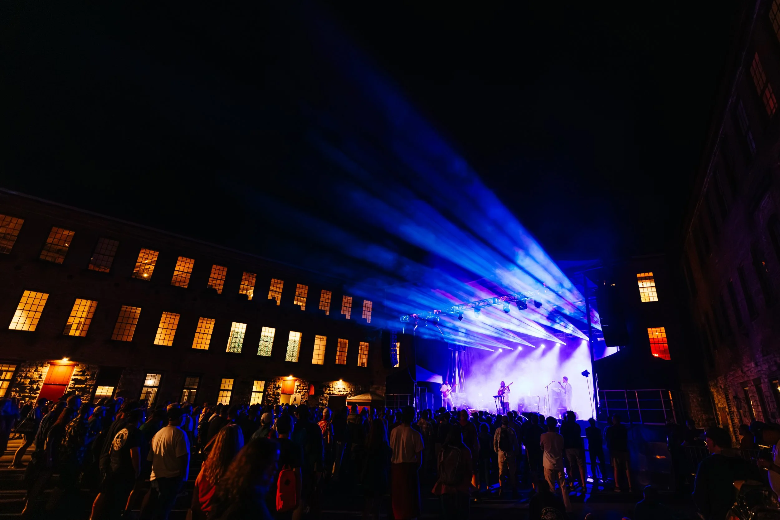 A large crowd of people gathered outdoors at night watching a live music concert with bright blue and purple stage lighting and a band performing.