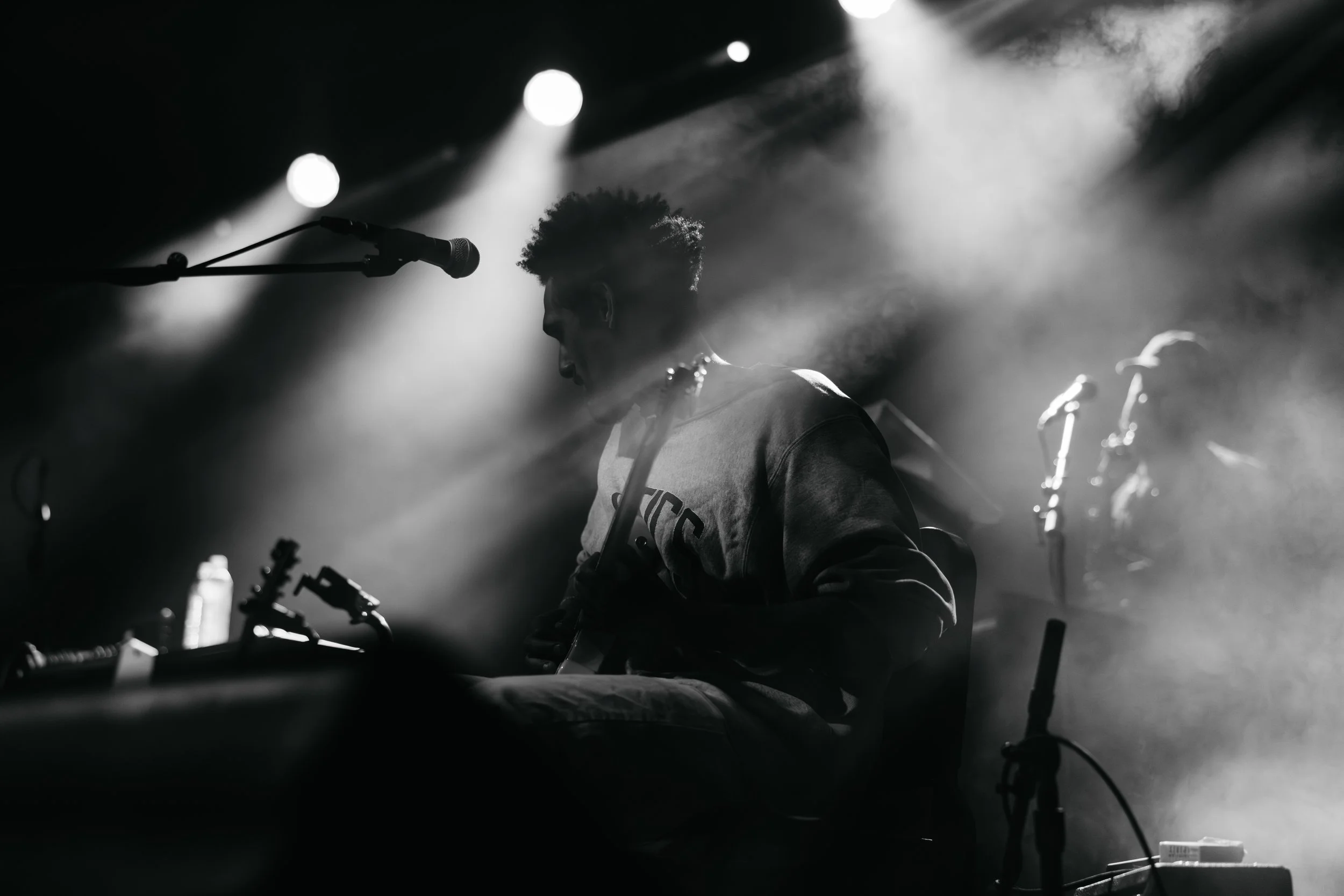 A black and white photo of a musician with curly hair playing a keyboard on a stage with dramatic lighting and smoke.