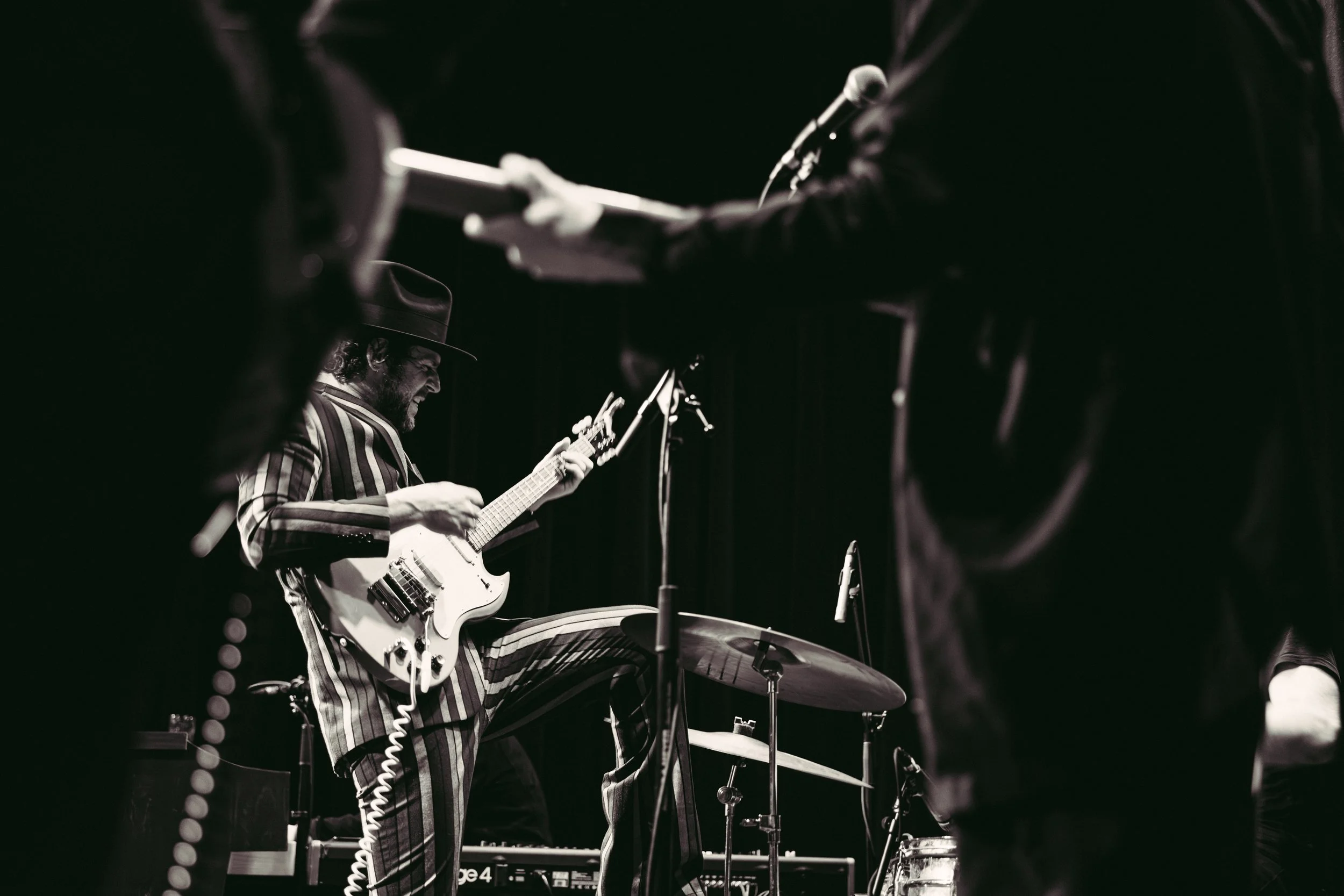 A musician wearing a striped suit and a hat playing an electric guitar on stage, with a microphone and drum set nearby, in a low-light performance setting.