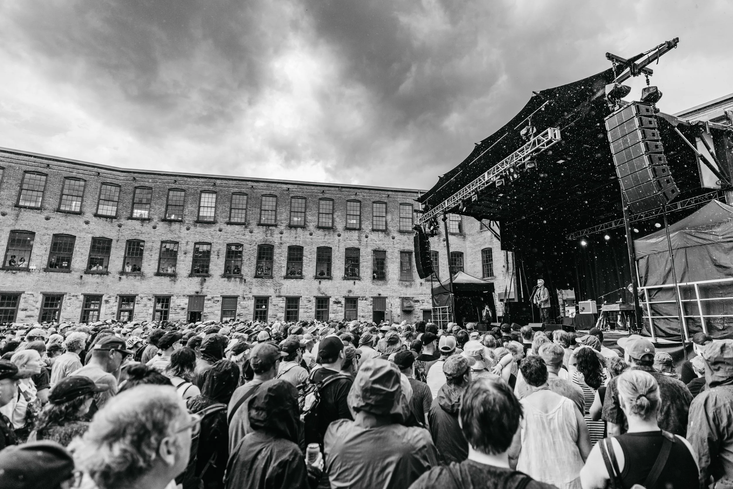 A large crowd gathered outdoors at a concert, facing a stage with musicians performing, in front of an industrial-style building with large windows, under a cloudy sky, all in black and white.