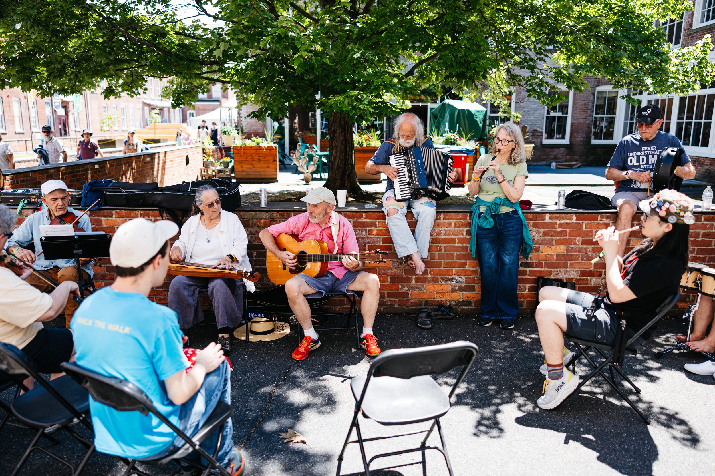 A group of older people playing musical instruments outdoors under a tree, with chairs and onlookers gathered around.
