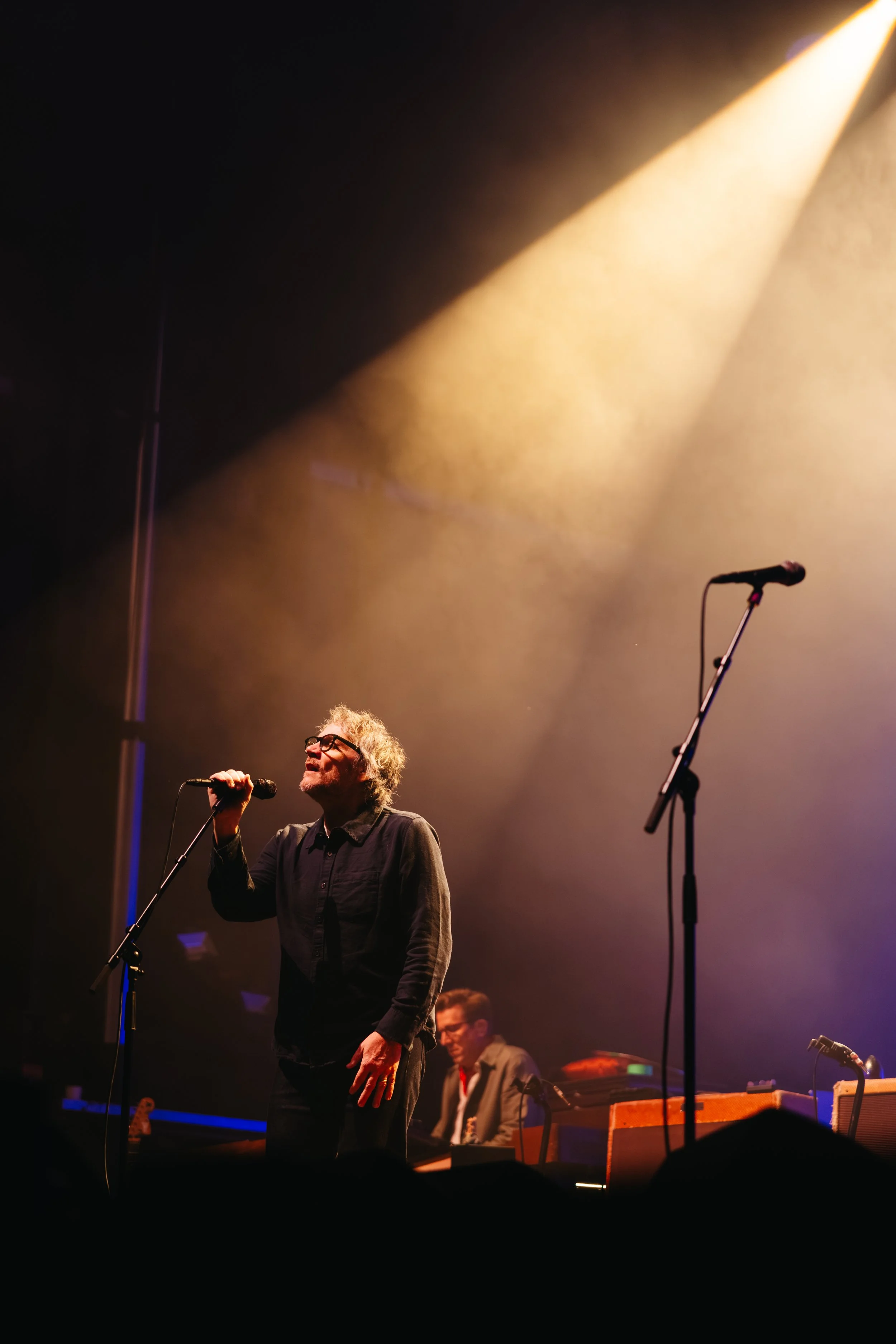 A man with curly, gray hair and glasses is singing into a microphone on a stage illuminated with yellow stage lights. There are musicians playing instruments in the background.