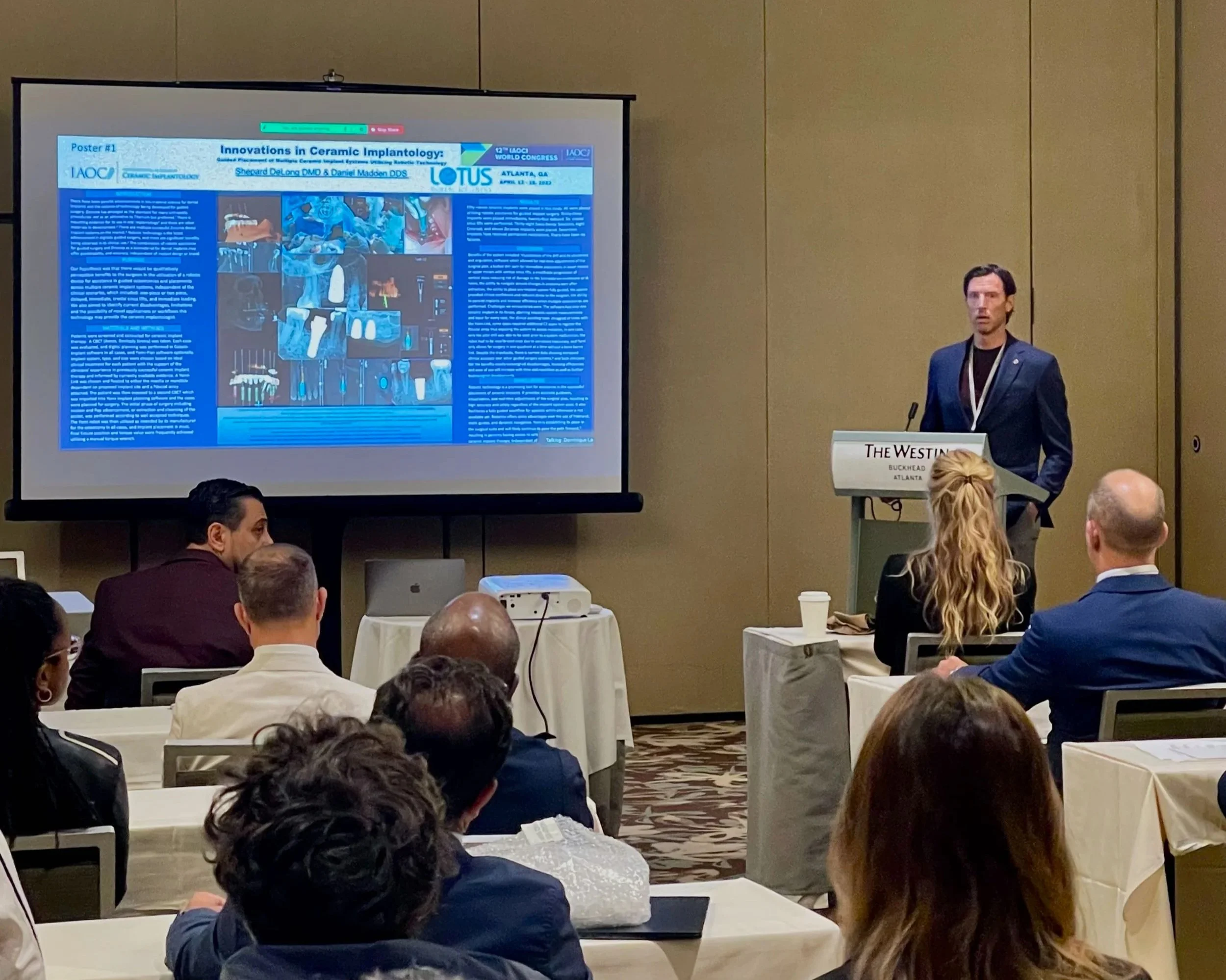 Conference room with a speaker at a podium presenting a research poster titled 'Innovations in Ceramic Implantology' to an audience. The presentation is projected on a large screen showing the poster with text and images of dental implants. The speaker is dressed in a dark suit and standing in front of a sign that reads 'The Westin Buckhead Atlanta.'
