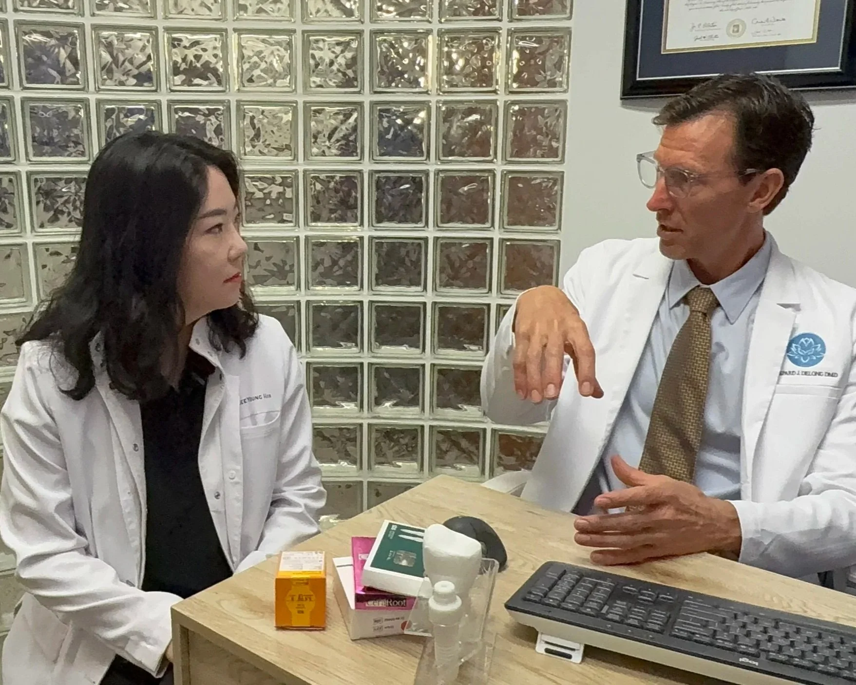 A male doctor and a female nurse or medical professional sitting at a desk in a medical office, engaged in conversation. The doctor is gesturing with his right hand, wearing glasses, a white coat, with a emblem on the coat pocket. The woman has dark hair, wearing a white coat and black top, listening attentively. The desk has medical supplies, pills, a keyboard, and a computer mouse. Glass blocks and framed certificate are in the background.