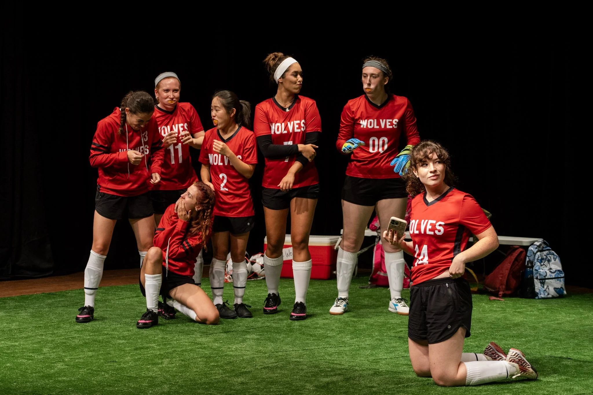 Group of young female soccer players wearing red jerseys with 'WOLVES' printed on them, standing on green turf, some with arms crossed or looking thoughtful, one kneeling on the ground.