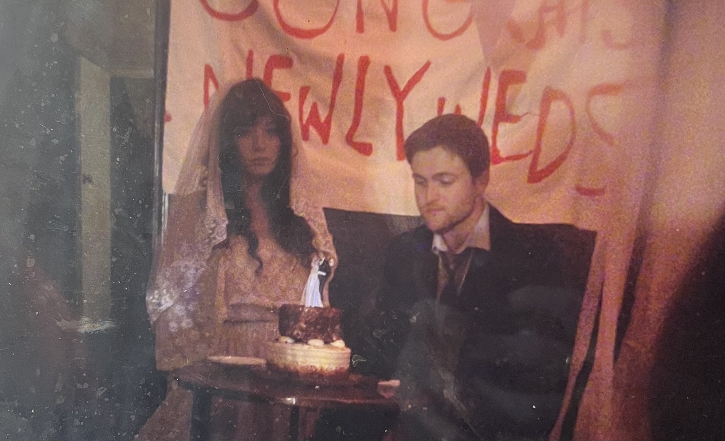 A man and a woman sit at a table with a birthday cake, in front of a banner with red writing, at a celebration. The woman is wearing a veil, the man has closed eyes.