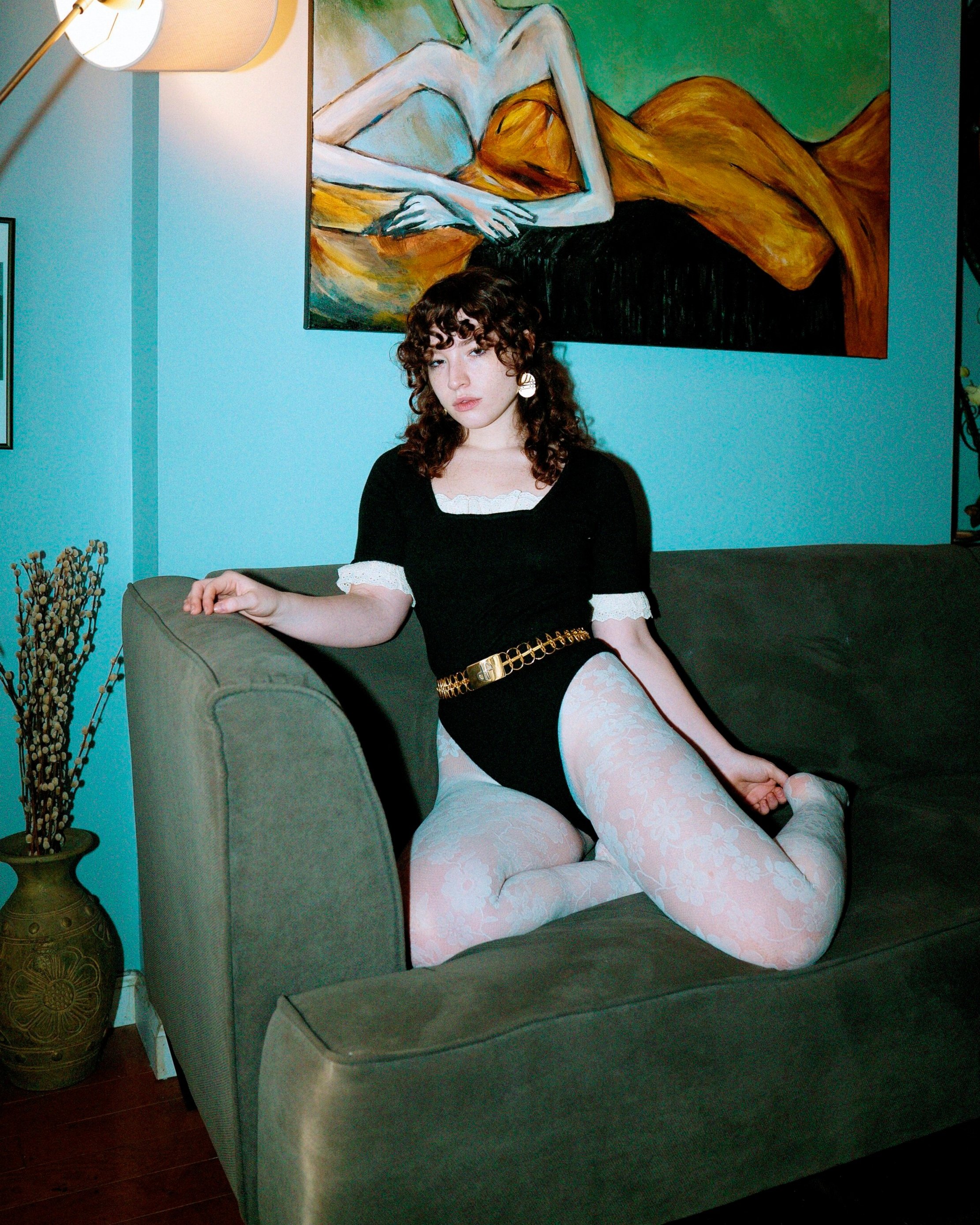 A young woman with curly brown hair and light skin poses on a green sofa in a room with teal walls, wearing a black and white dress and floral tights, with a large colorful painting of a reclining figure hanging behind her.