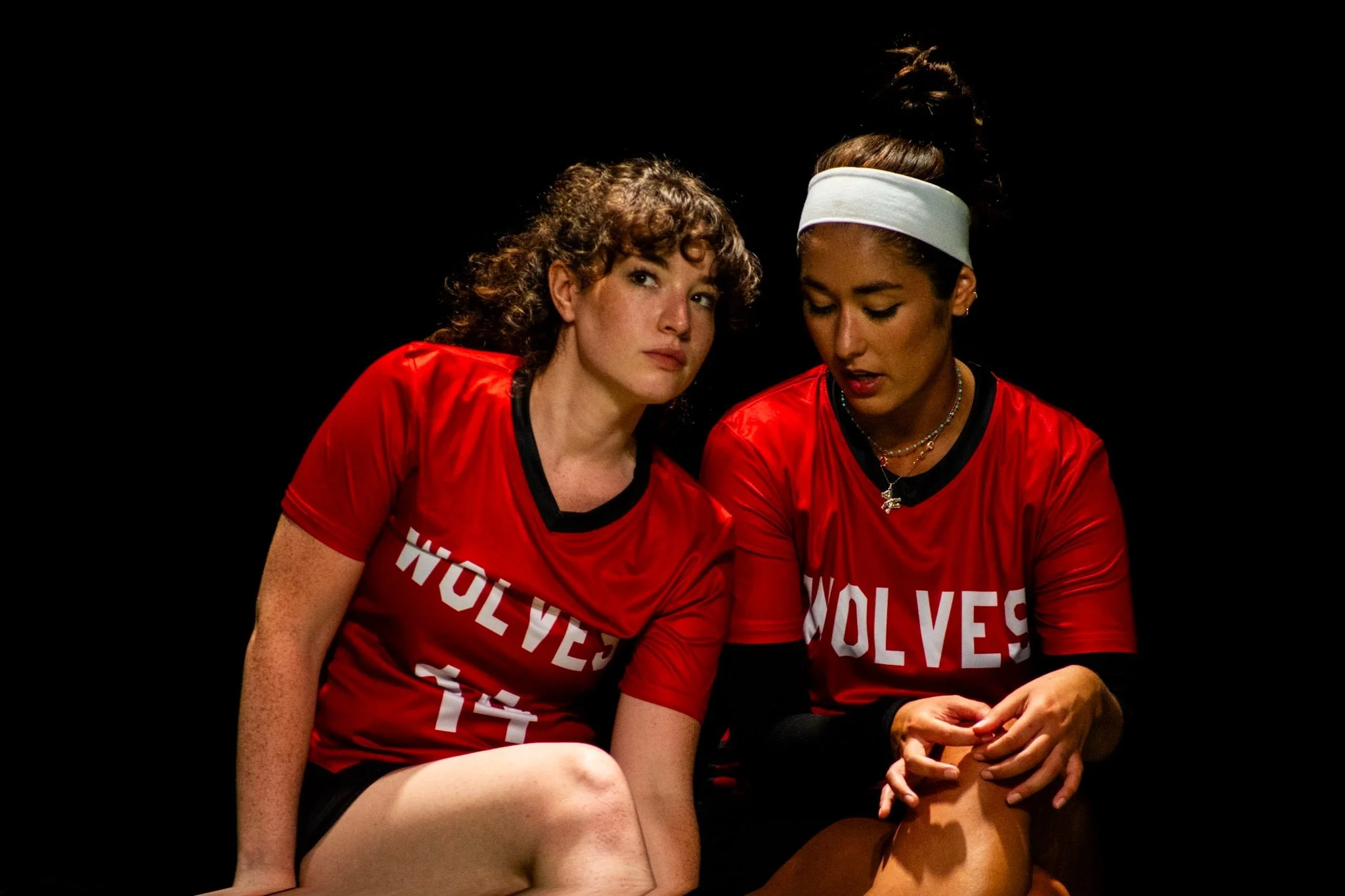 Two female athletes in red 'Wolves' jerseys sitting together against a black background, one with curly hair and the other with a headband, looking down and to their sides.