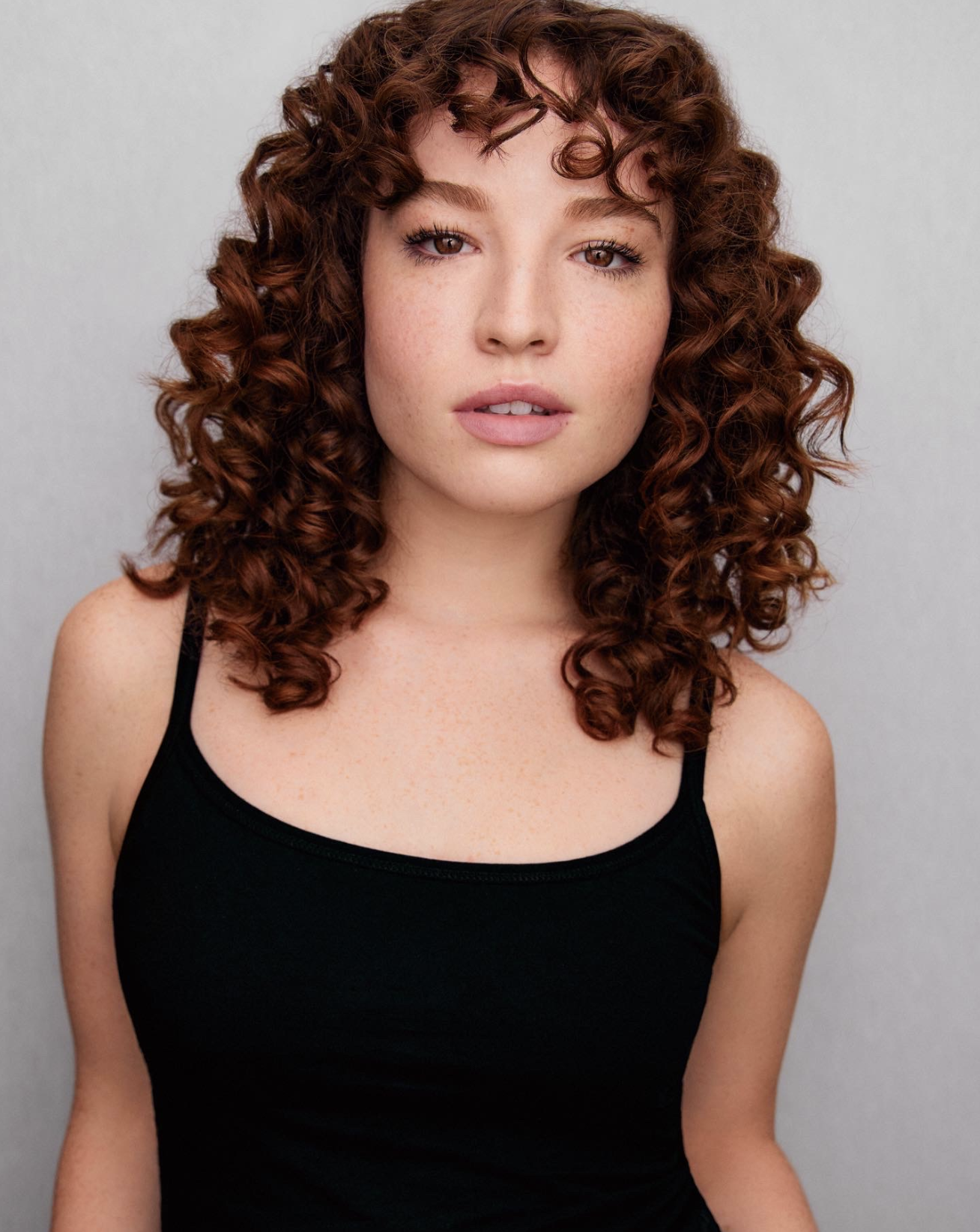 A young woman with curly red hair and freckles wearing a black tank top, looking directly at the camera.