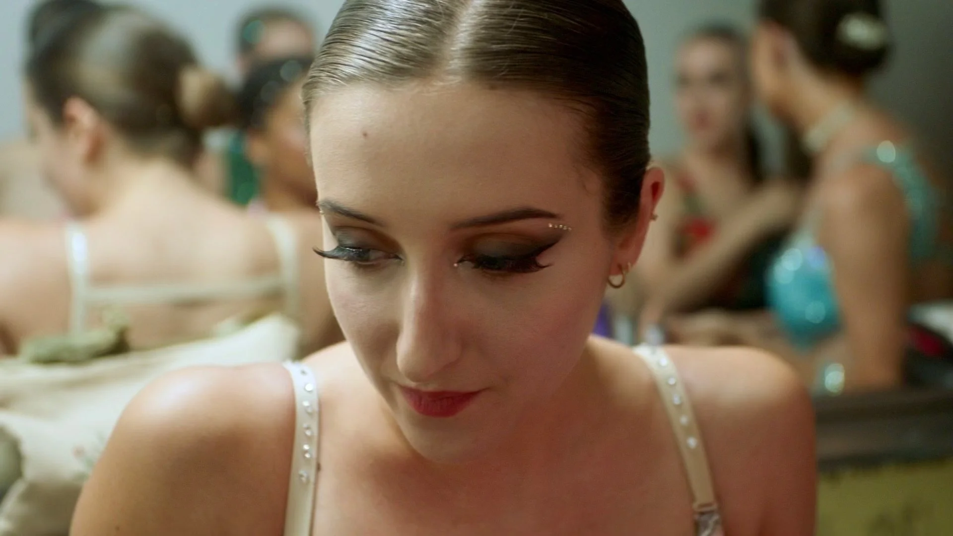 A young woman with brunette hair tied back, wearing makeup with dark eyeshadow, false eyelashes, and a pearl-studded top, is looking down in a dressing room with other women preparing in the background.