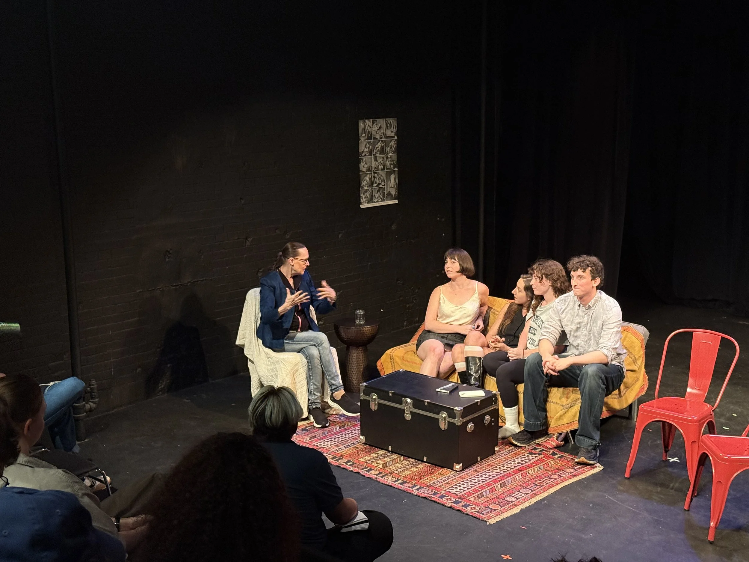 A group of five people sitting on a sofa and chairs on stage, engaged in a conversation during a theatrical performance, with an audience watching.