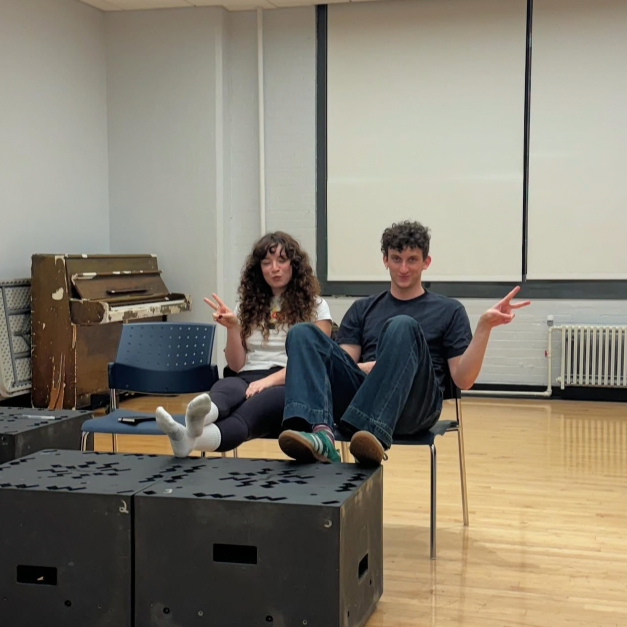 Two young people sitting on chairs in a room, making peace signs with their hands, with a piano and a large blank whiteboard behind them.