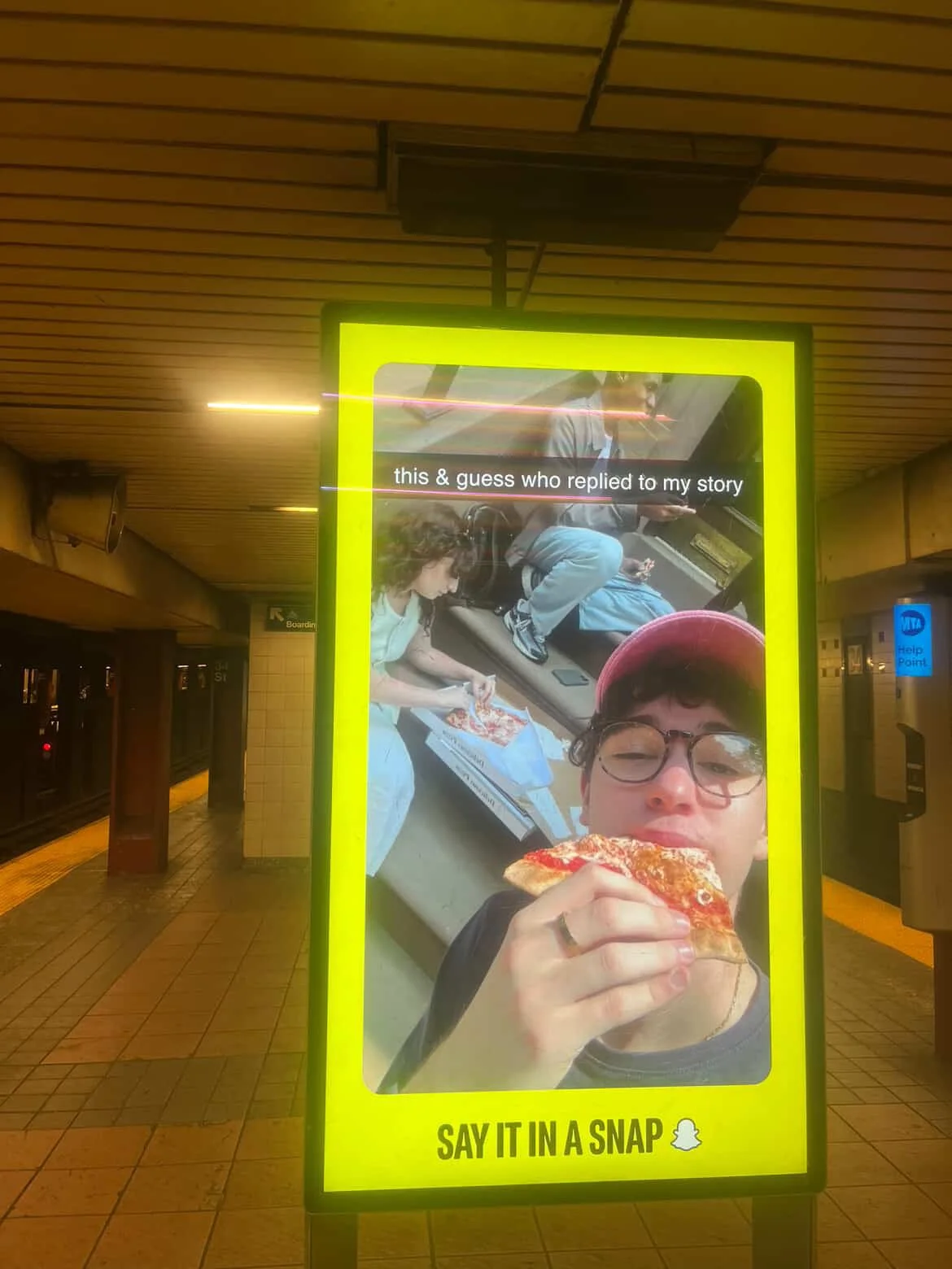 Bright yellow sign with a photo of a person wearing glasses and a red hat eating pizza, and another person and a child in the background. Text says, 'this & guess who replied to my story' and 'SAY IT IN A SNAP' with Snapchat logo.