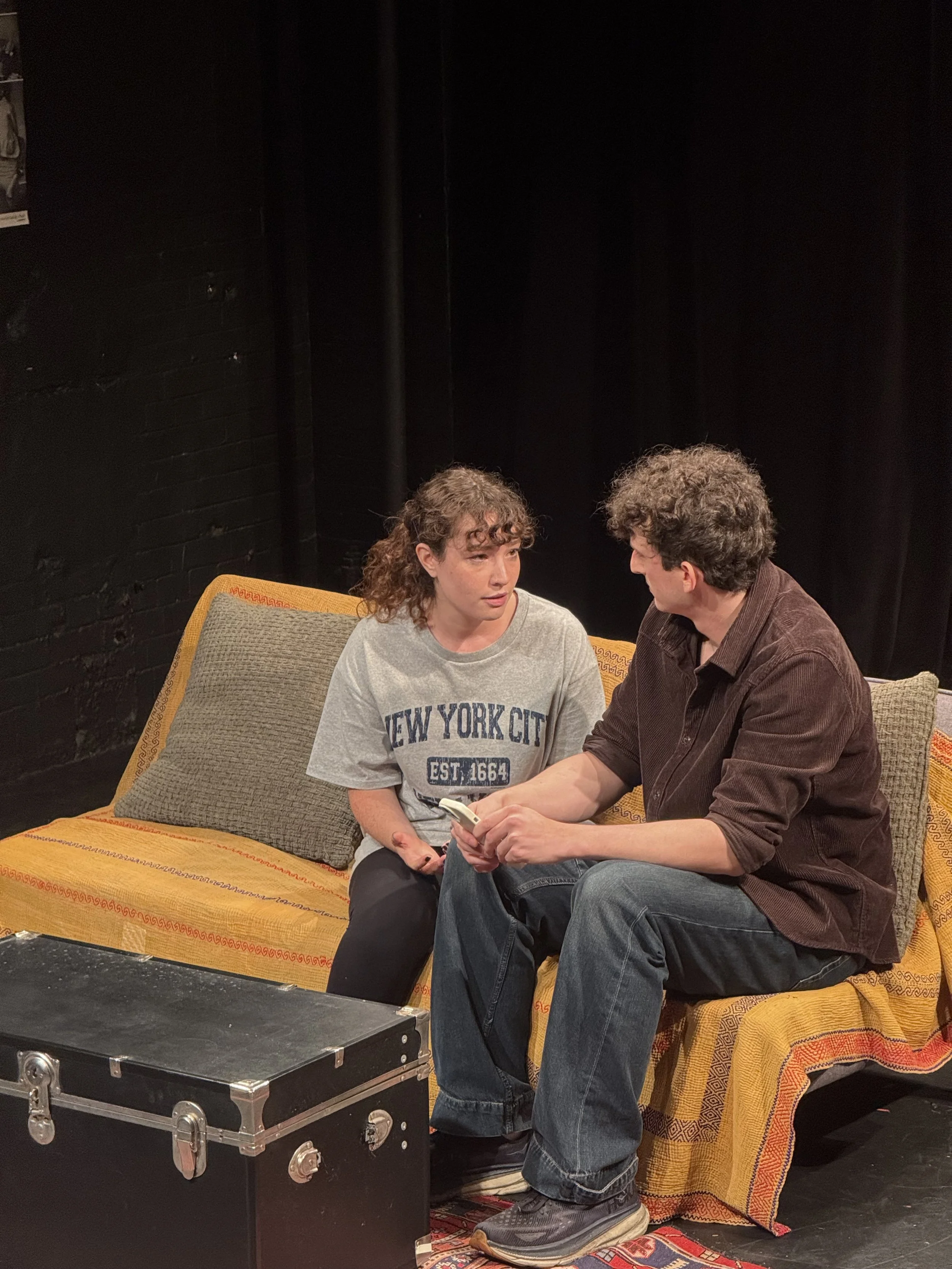 A young woman and a young man sitting on a yellow couch, engaged in conversation. The woman is wearing a gray 'New York City' t-shirt, and the man is holding a remote control, wearing a brown shirt and jeans. They are on a stage with a black backdrop, and a black storage box is in front of them.