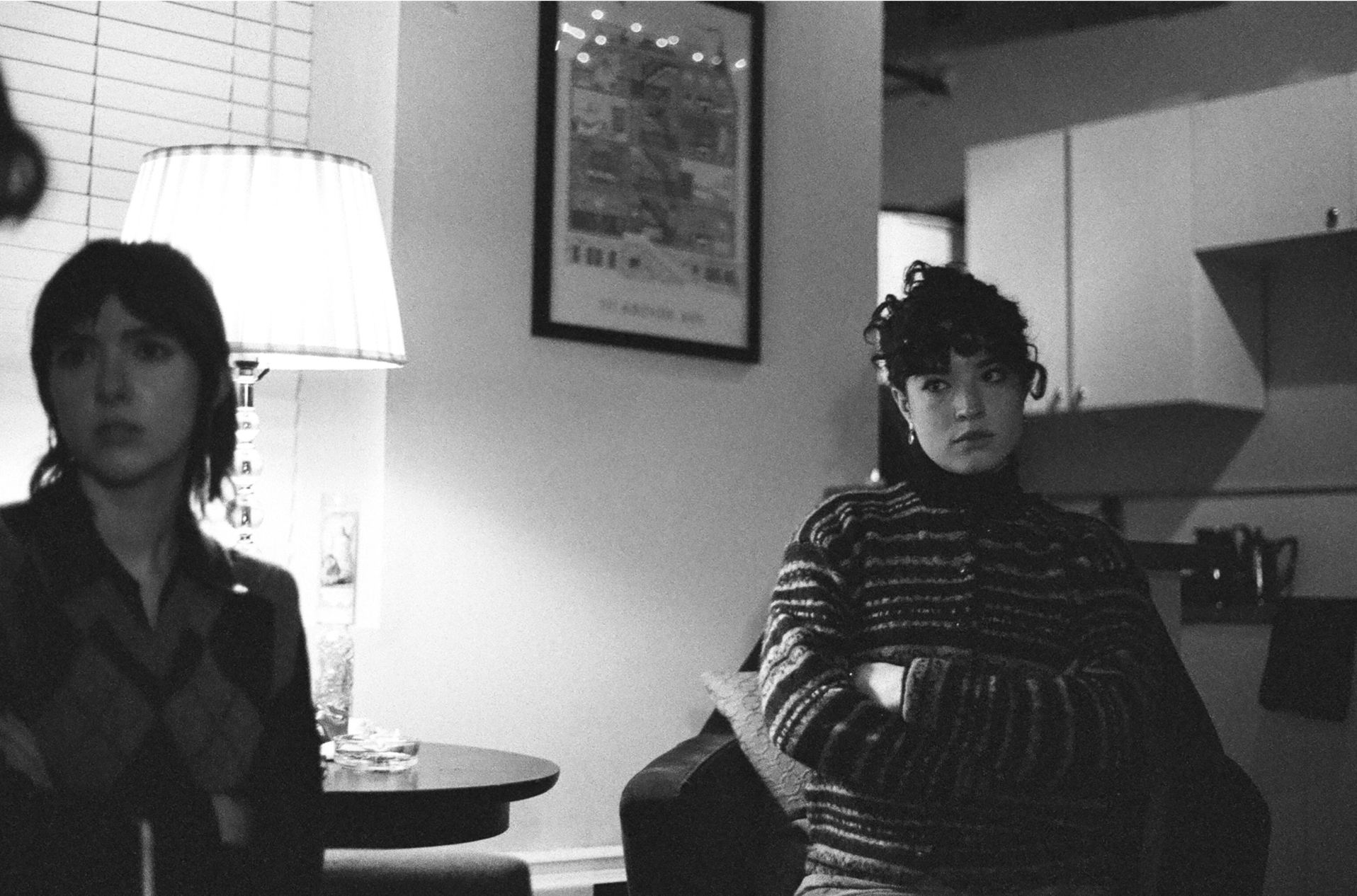 Two women sitting in a dimly lit room, one near a lamp, with a painting on the wall and kitchen cabinets in the background.