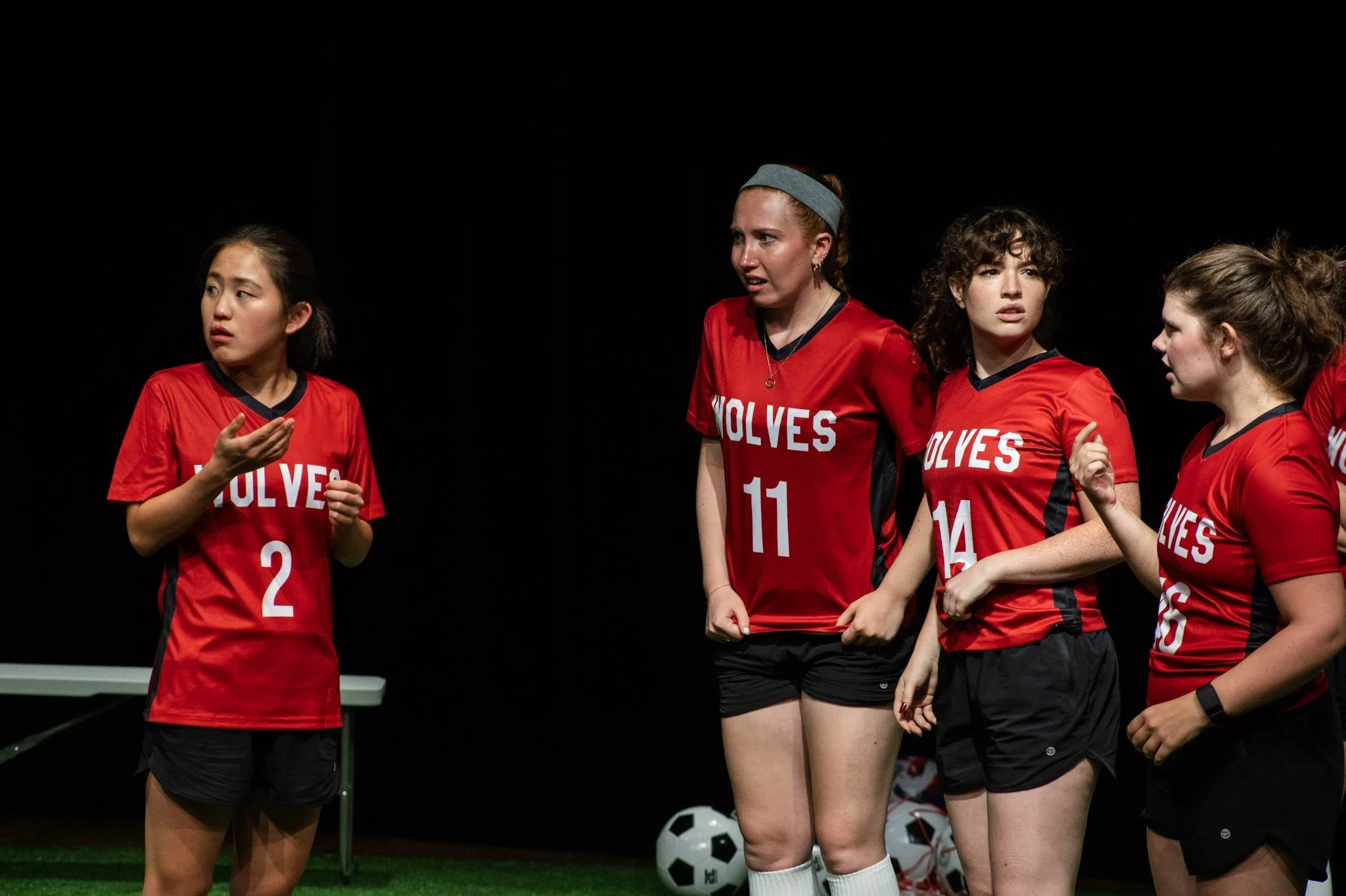 Four young women in red soccer jerseys with 'WOLVES' and numbers, standing on an indoor field, engaged in a conversation or discussion.