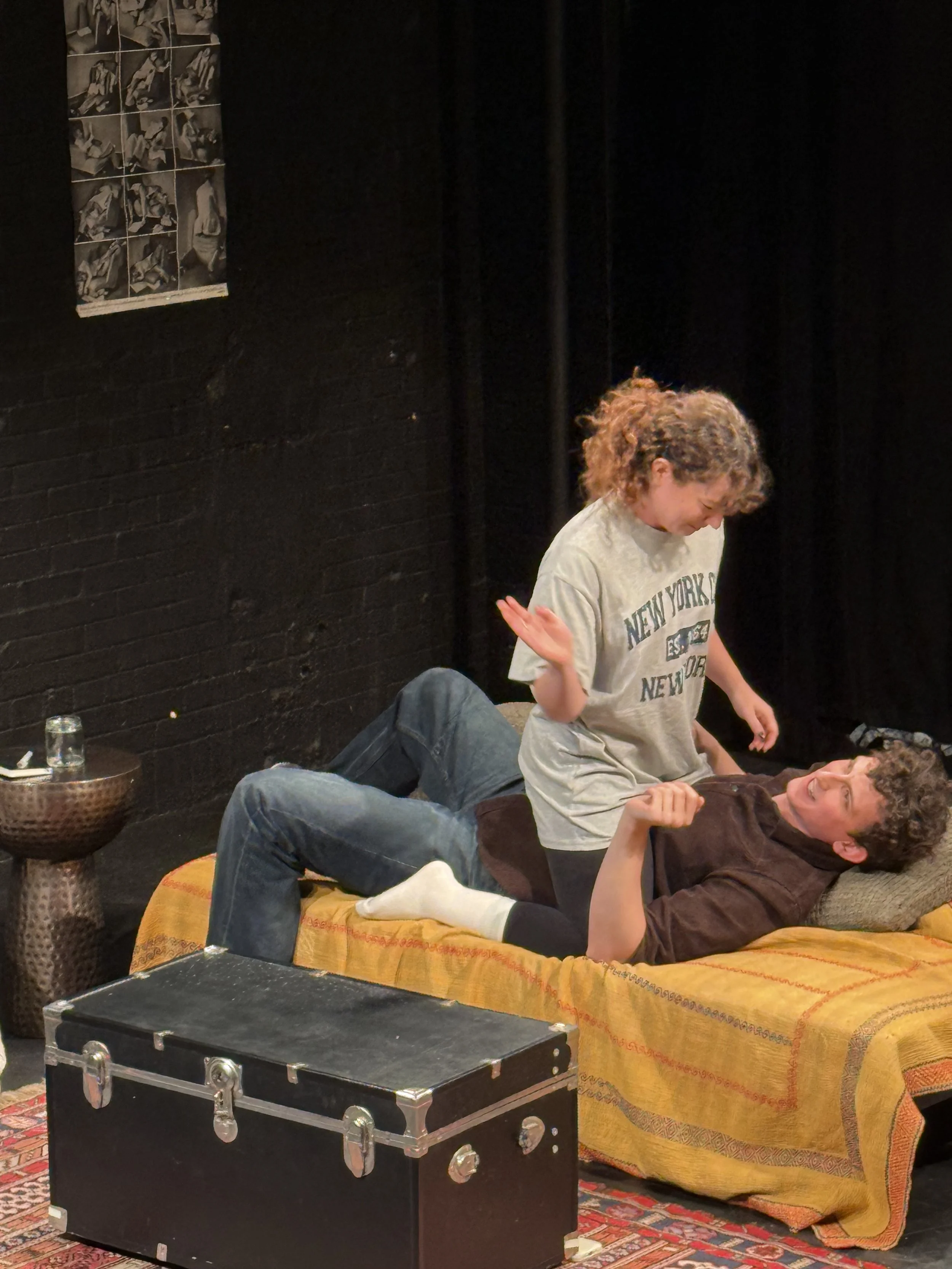 Two young women with curly hair laughing and engaging in a playful moment on a sofa with a yellow blanket, surrounded by stage props including a black trunk, a small side table with a glass, and a poster on a black wall in a theater or performance sp