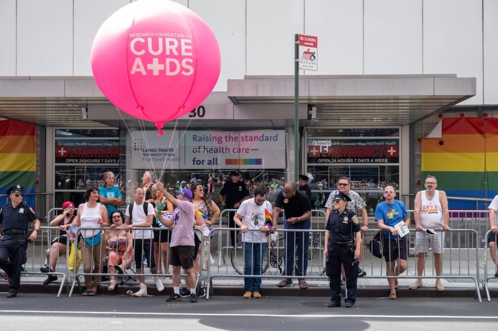 People gathered outside a healthcare facility holding rainbow flags and balloons with a pink balloon promoting research to cure AIDS, during a Pride event with police officers present.