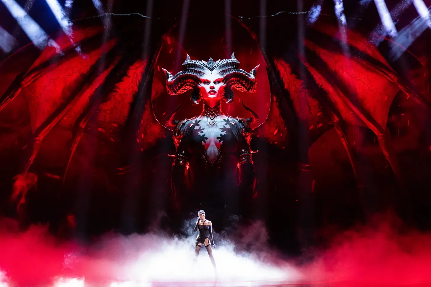A performer standing on stage with dramatic red lighting, fog effects, and an elaborate backdrop depicting a demonic character with horns, wings, and a fierce expression.