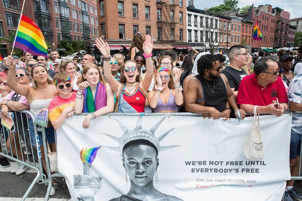Crowd of diverse people at a pride parade, some holding rainbow flags, behind a banner with a drawing of the Statue of Liberty wearing a crown that resembles a flame, advocating for AIDS awareness.