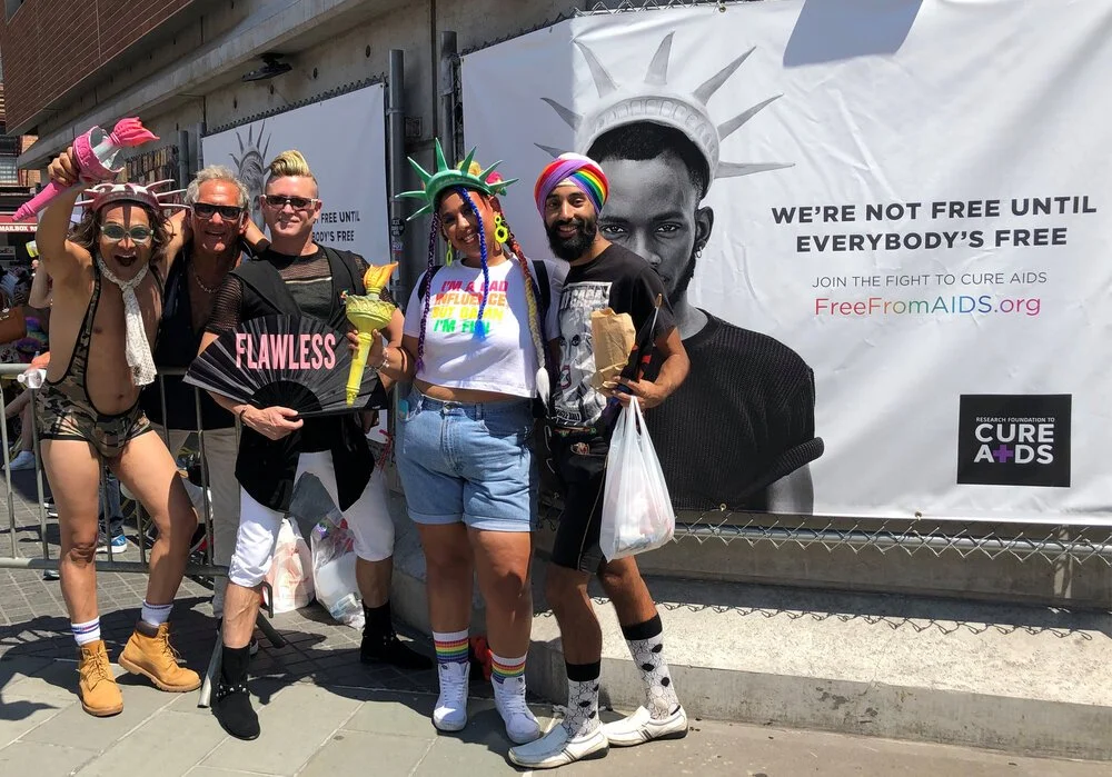 Group of five people dressed in colorful, playful costumes at an AIDS awareness event. They are standing in front of a banner promoting AIDS awareness and cure. They are smiling, some holding props and wearing rainbow accessories.