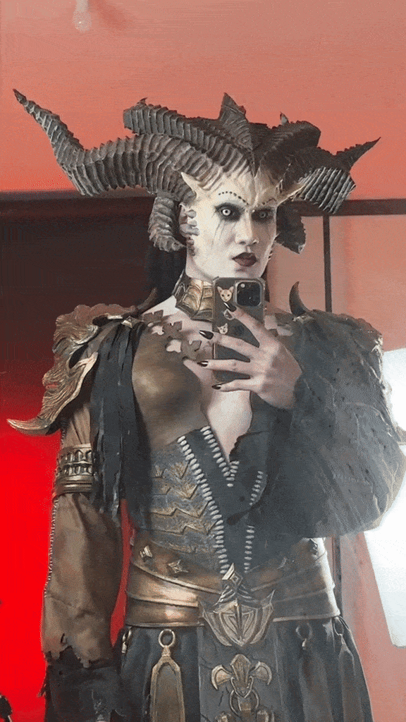 Person dressed as a mythical or fantasy character with large horned headdress, elaborate armor, dark makeup, and black nail polish, taking a selfie in a mirror.