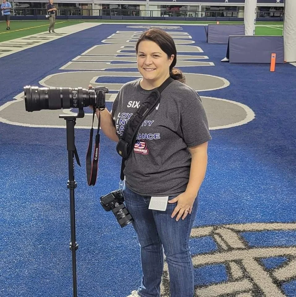A woman standing on a blue football field with a large camera lens on a tripod, holding a camera, smiling at the camera. She is wearing a gray T-shirt and jeans, with a smaller camera hanging from her neck. The football field has painted yard lines and a padded goal post in the background.
